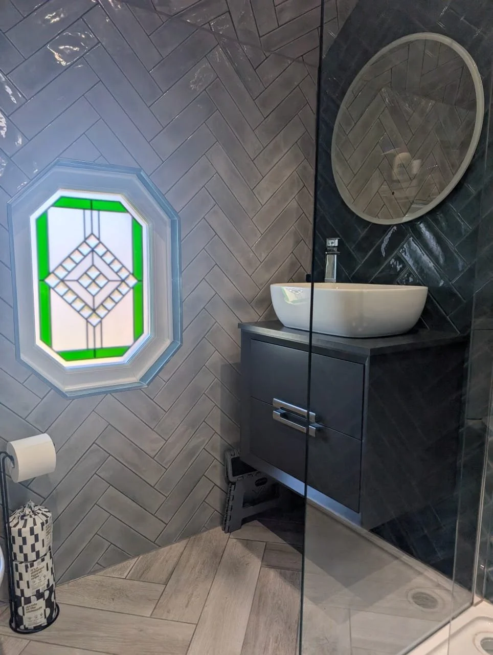 A modern bathroom with herringbone patterned gray tile walls and a window with a stained glass design. There is a dark gray vanity with a white vessel sink, a round mirror on the wall, and a black step stool on the floor. Toilet paper and a toilet pa