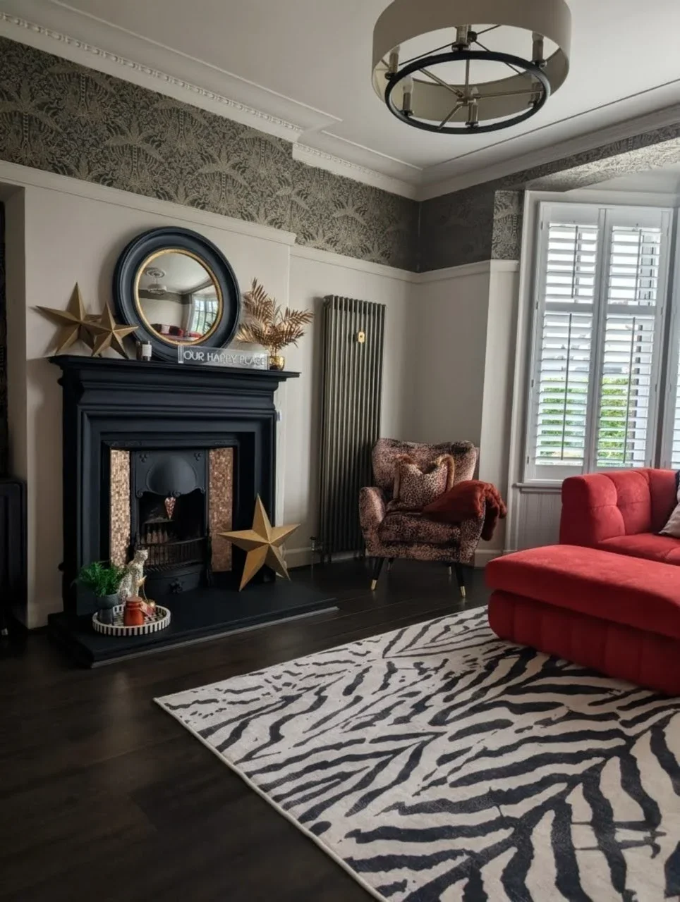 Living room with a fireplace, decorated with gold stars and a mirror, a zebra print rug on dark wood floor, red sofa, patterned armchair, window with white shutters, and modern ceiling light.