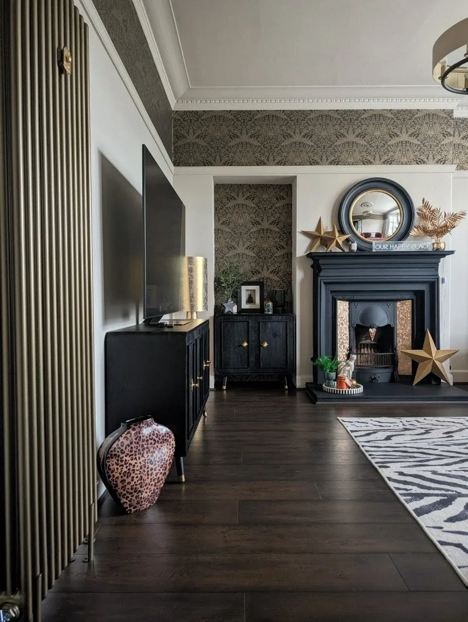 Living room with dark hardwood floor, black fireplace surrounded by gold and black decor, leopard print vase, zebra print rug, black cabinet, flat-screen TV, gold lamp, wall with decorative wallpaper, and various decorative items including gold stars, plants, and a mirror.