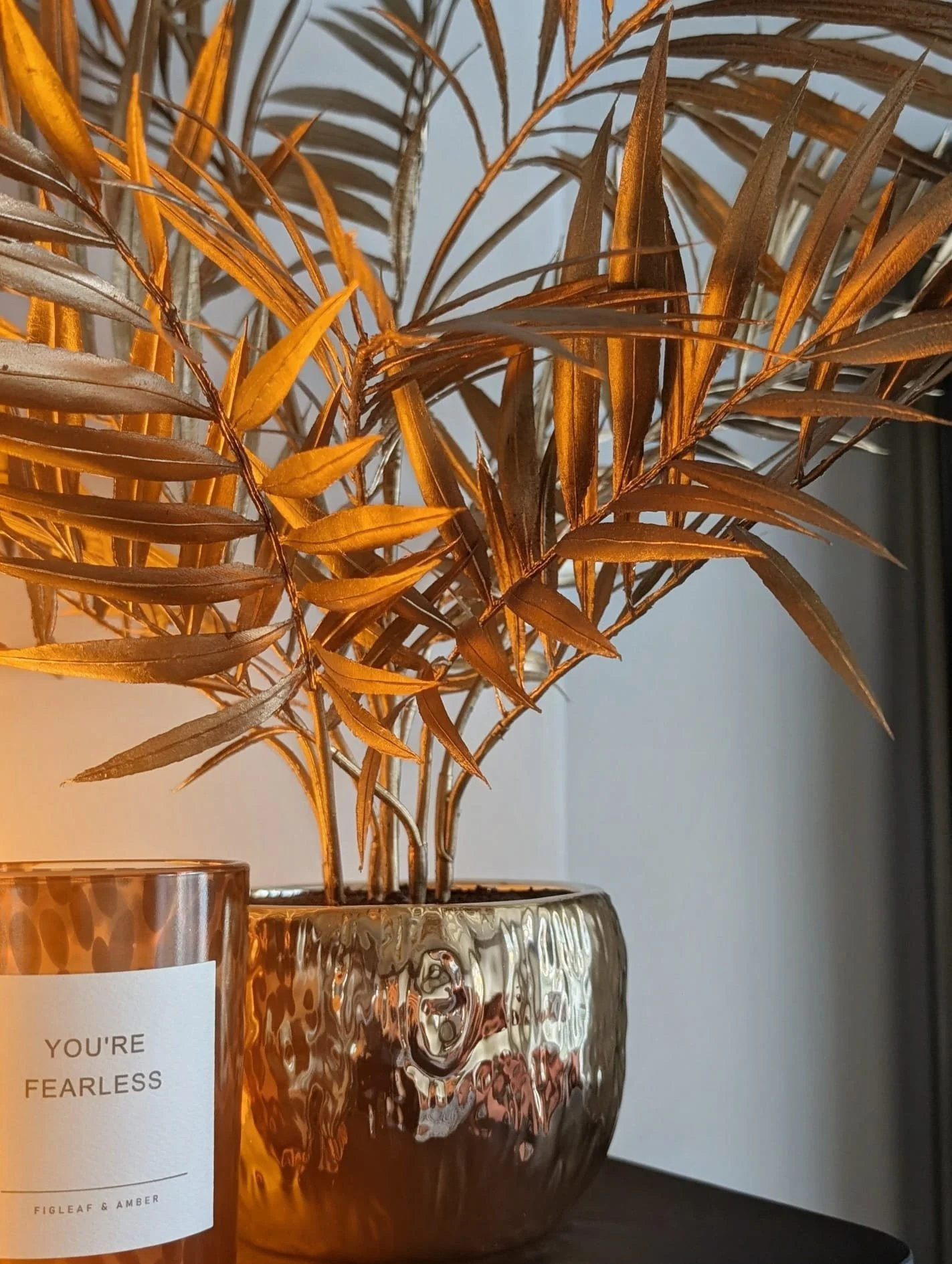A decorative metallic gold vase with an embossed pattern holding dried orange-brown leaves, placed on a dark surface against a neutral background.