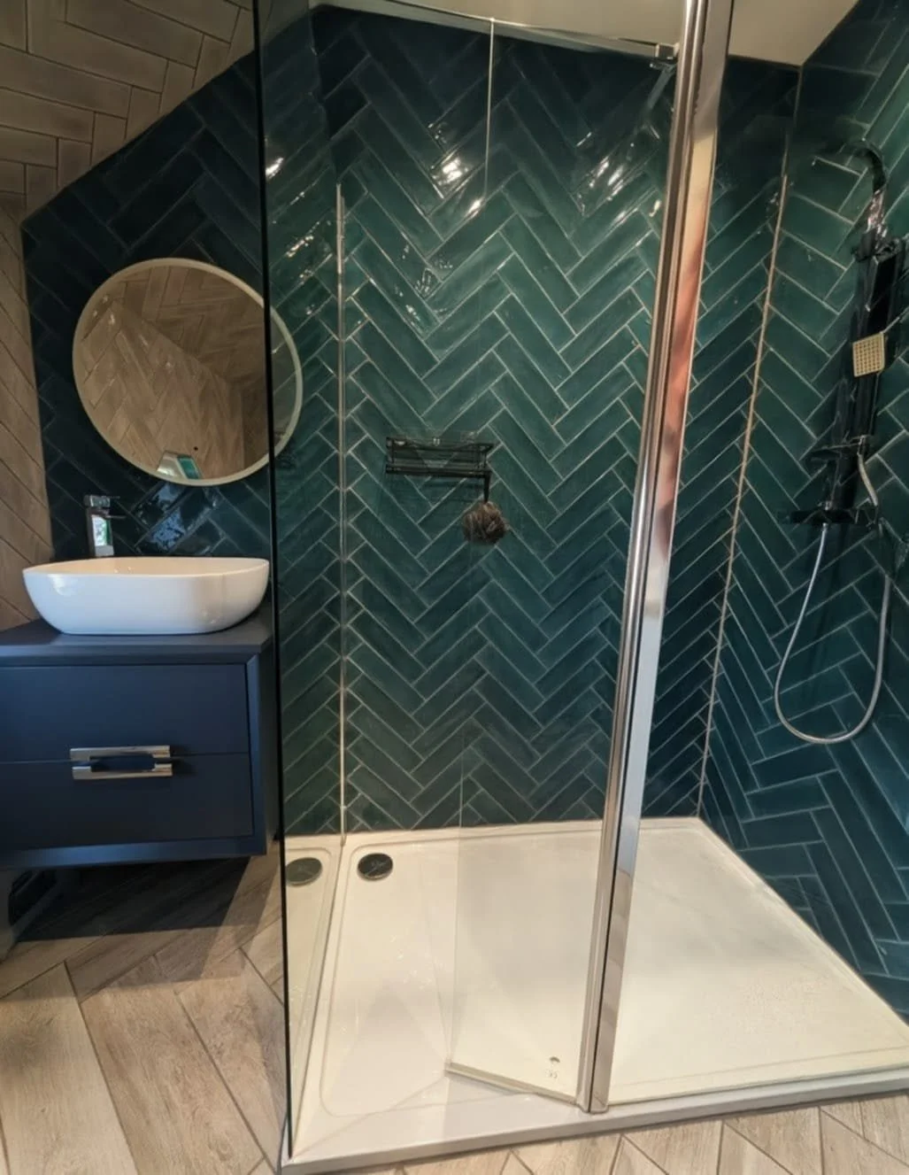 Modern bathroom with a walk-in shower featuring dark blue herringbone tiles, a glass door, a wall-mounted showerhead and handheld shower, a mirror, and a blue vanity with a vessel sink.