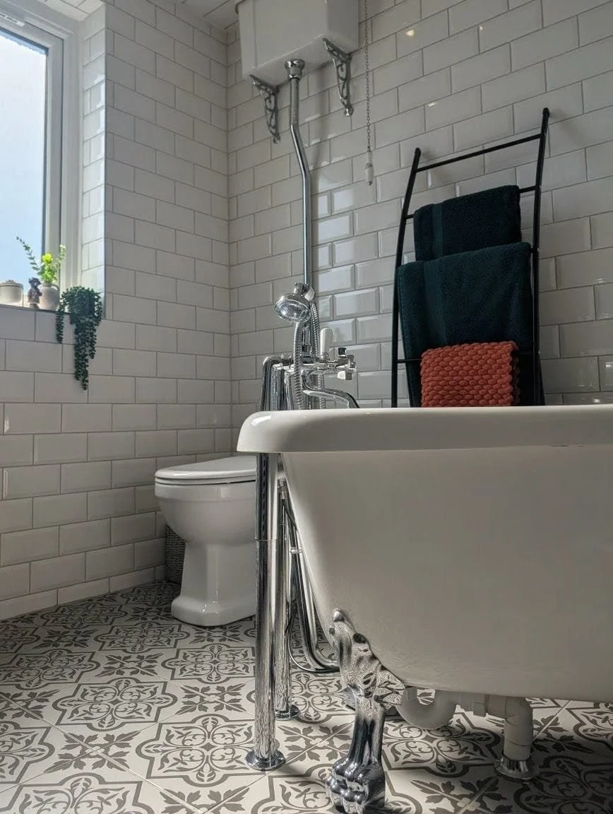 A bathroom with white subway tile walls, a window with small potted plants, a vintage clawfoot bathtub, a modern toilet, and open shelves with towels. The bathroom floor has patterned tile.