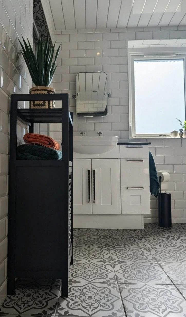 Bathroom with white subway tile walls, tiled floor with intricate black and white pattern, black shelving unit with folded towels, white vanity with a round white sink, small mirror above sink, window allowing natural light, potted plant on window si