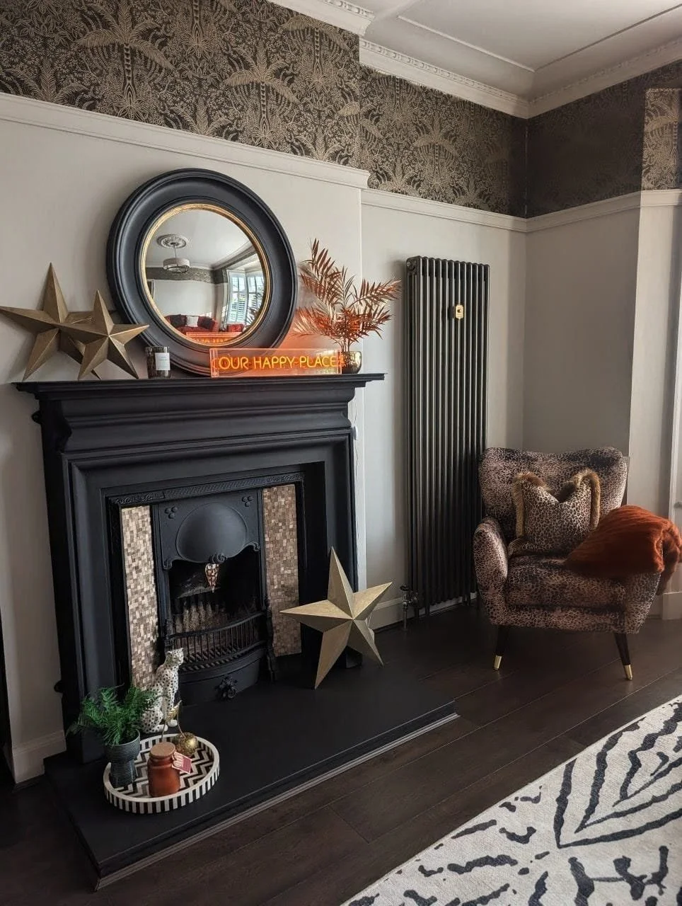 Living room corner with a black fireplace, round mirror, gold star decorations, a neon sign reading 'Our Happy Place,' a leopard-print armchair with an orange pillow and throw, and a white and black patterned rug.