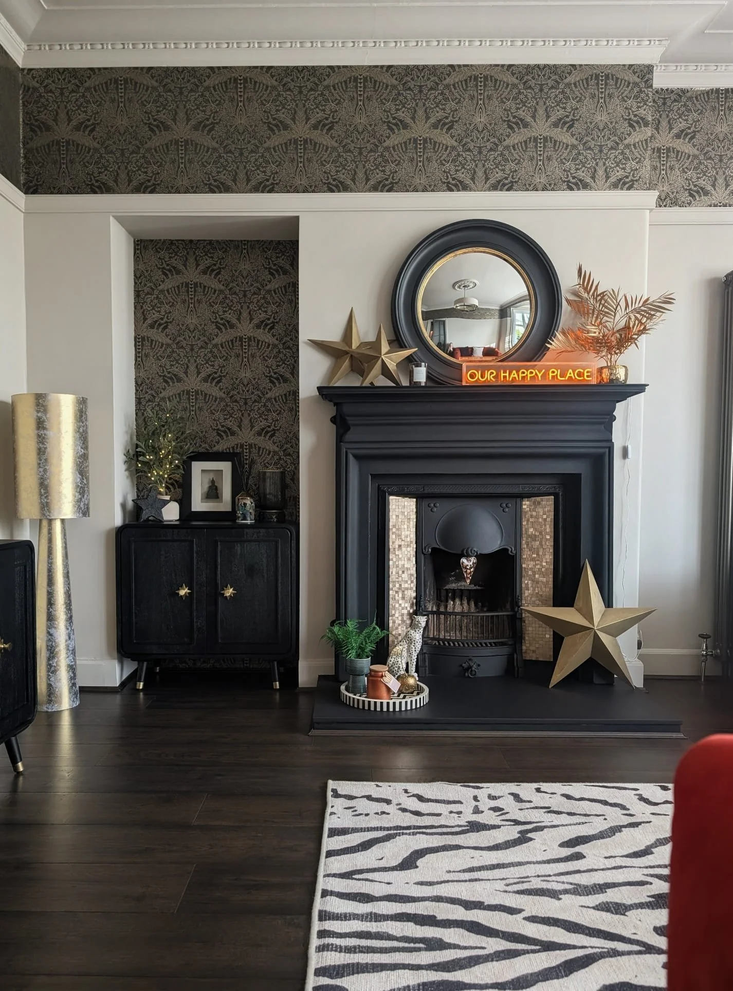 A living room fireplace with vintage black mantel, decorated with gold star ornaments, a round mirror, and a neon sign stating "Our Happy Place." Surrounding decor includes a large gold star, a potted fern, small animal figurines, and a striped tray 