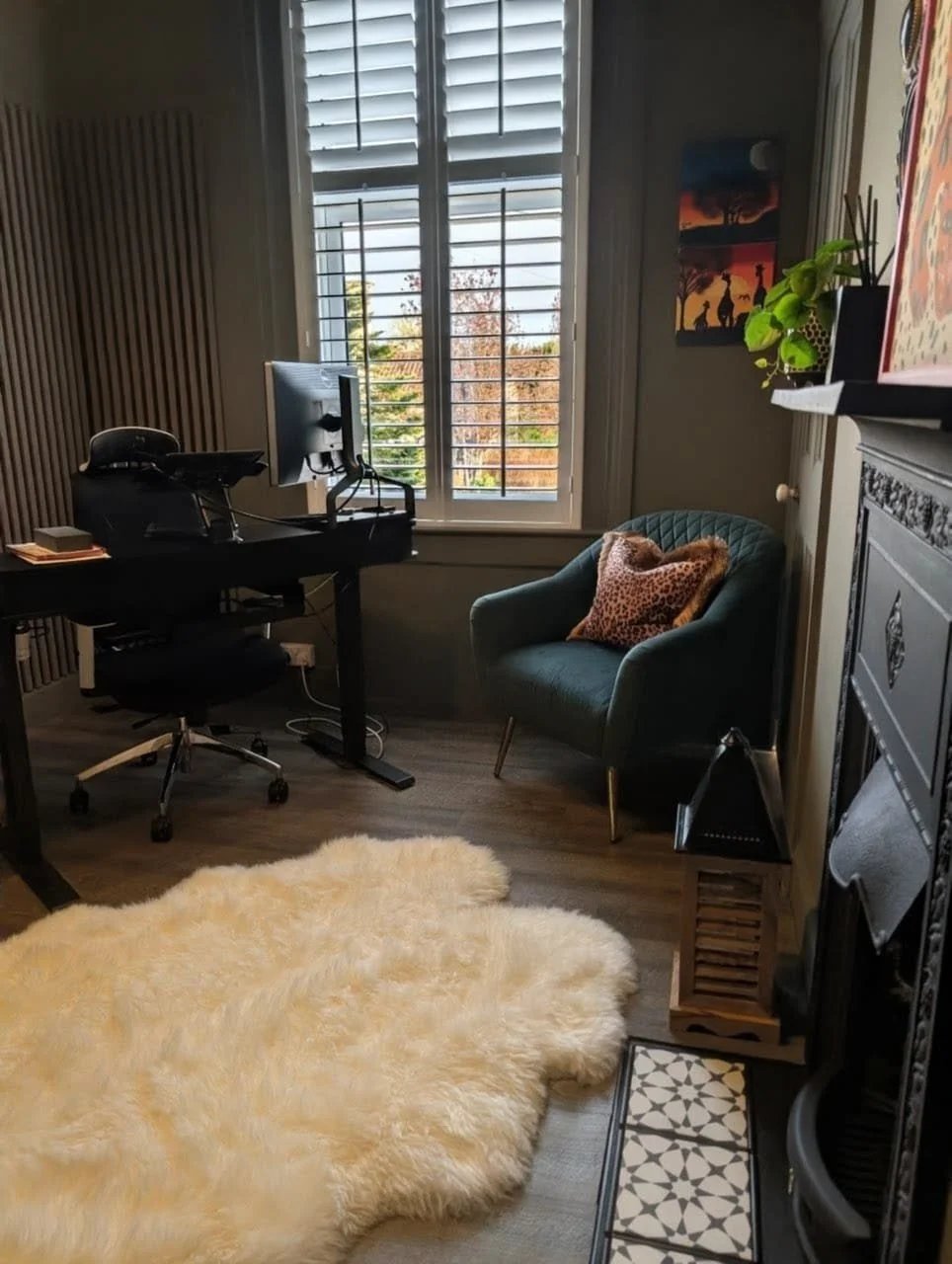 A cozy home office with a black desk and computer, an office chair, a green armchair with a leopard print pillow, a cream fluffy rug, and a window with shutters showing autumn trees outside.