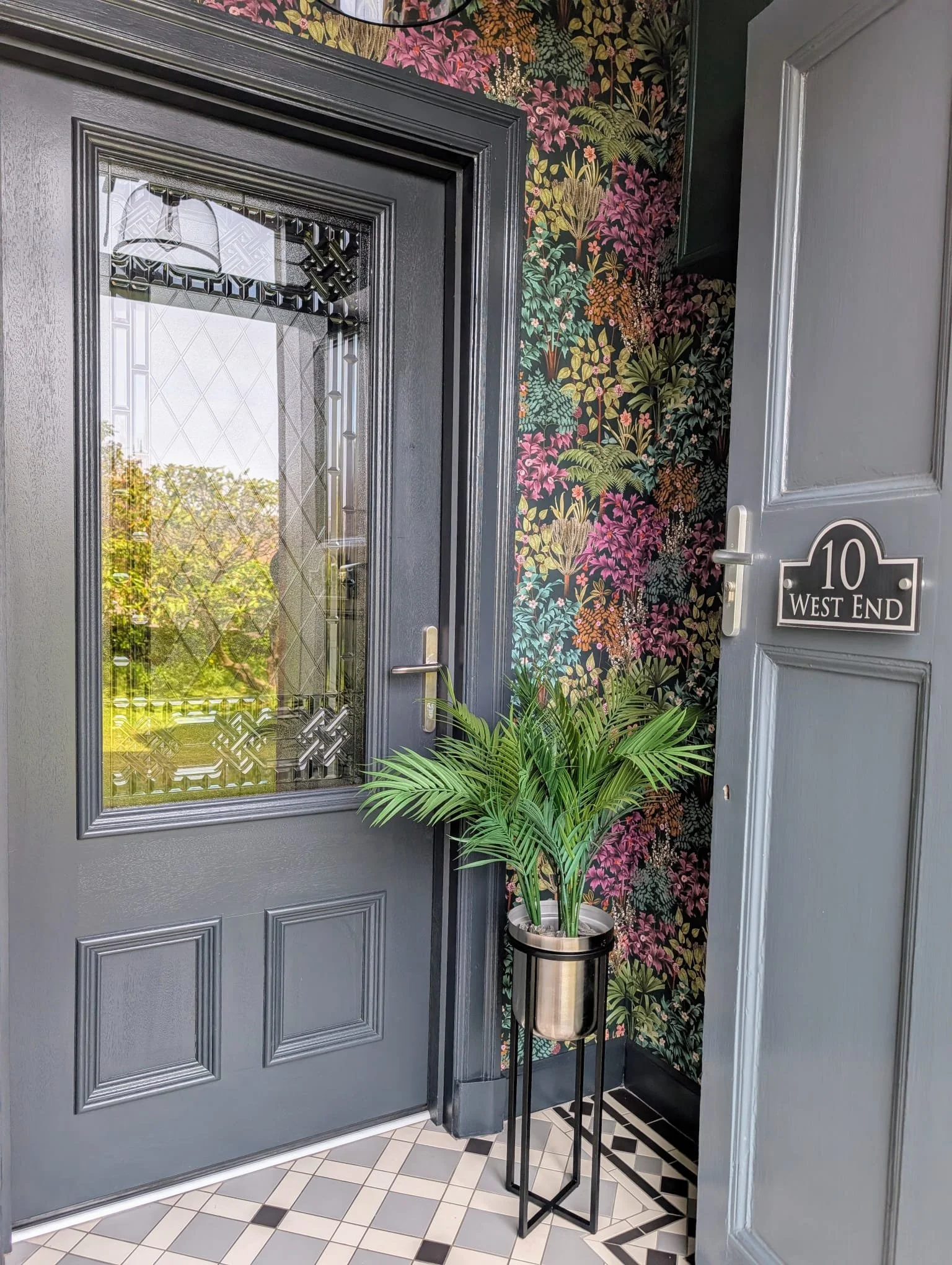 A gray front door with decorative glass window, black framing, and a handle, set in a house with black and pink floral wallpaper behind it. To the right, there is a gray wall and a door with house number 10 and the words 'West End'. A potted green pl