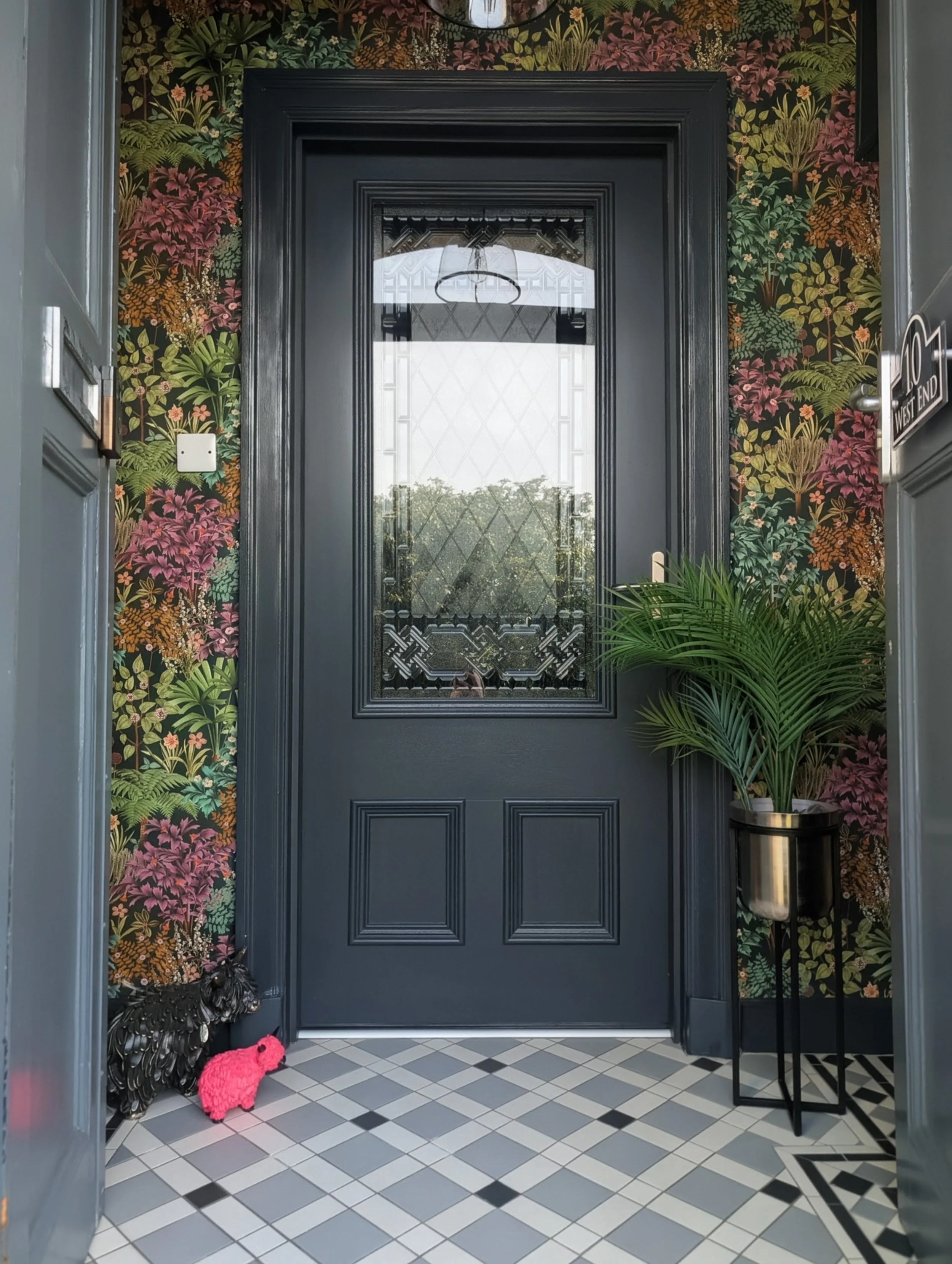 The image shows a black front door with a large glass window, surrounded by floral wallpaper in dark green, pink, orange, and yellow tones. There are decorative plants in black and metallic pots, and a pink dog-shaped toy on the floor. The floor is tiled in a black and white geometric pattern, and a small black side table is placed beside the door. A sign on the door reads 'West End'.