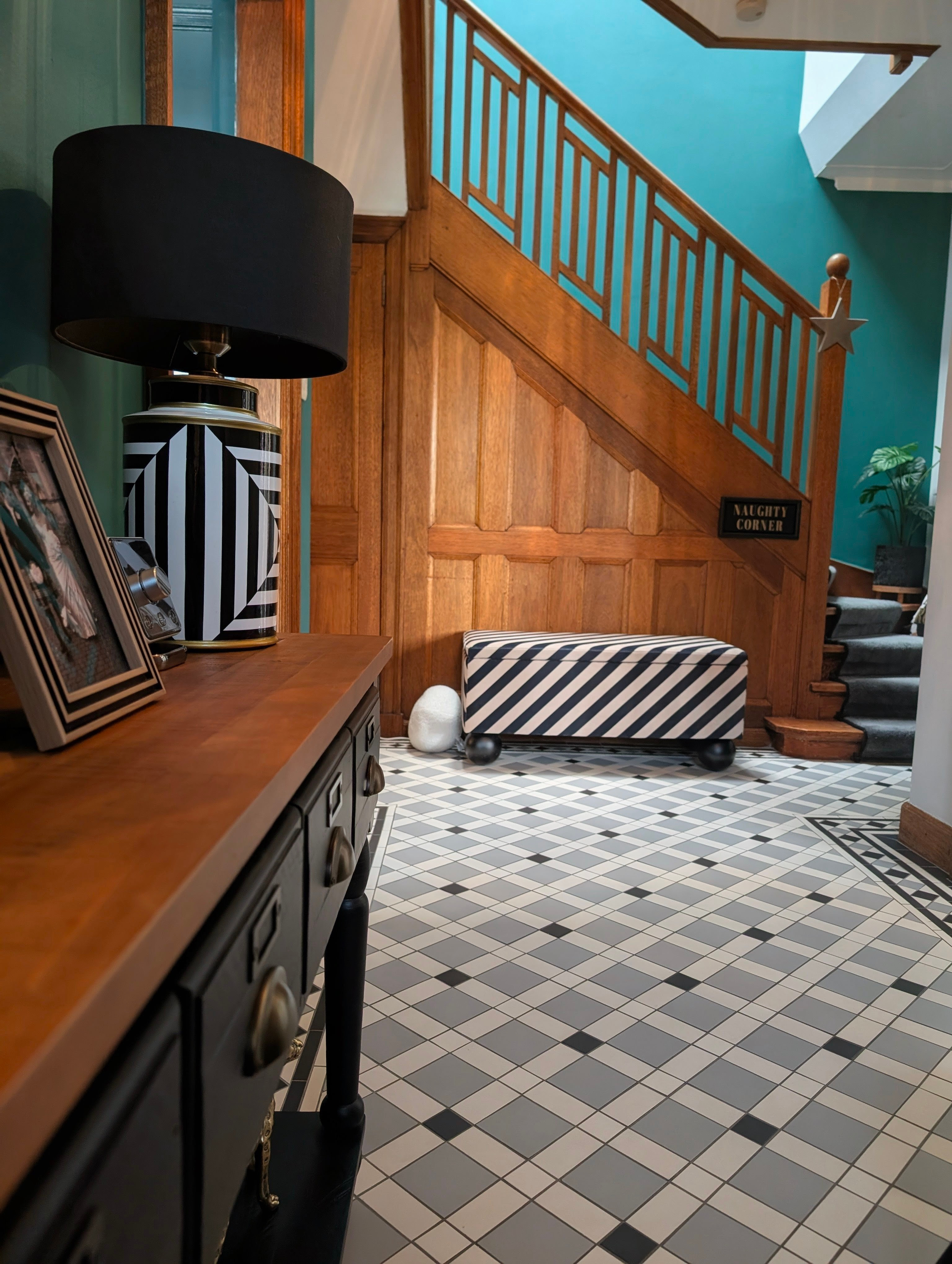 Interior view of a room with a black and white striped table, a wooden cabinet, a black lamp with a shade, framed photos or artwork, a small decorative object, and a patterned floor. A staircase with wooden railing leads upstairs, with a sign reading