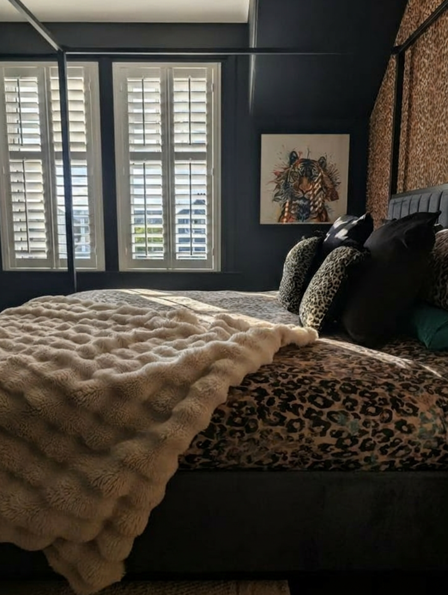 A bedroom with a bed covered in a textured beige blanket and decorative pillows with animal print, black, and beige designs. There are leopard print curtains and a framed painting of a tiger on the dark-colored wall. Large windows with white shutters