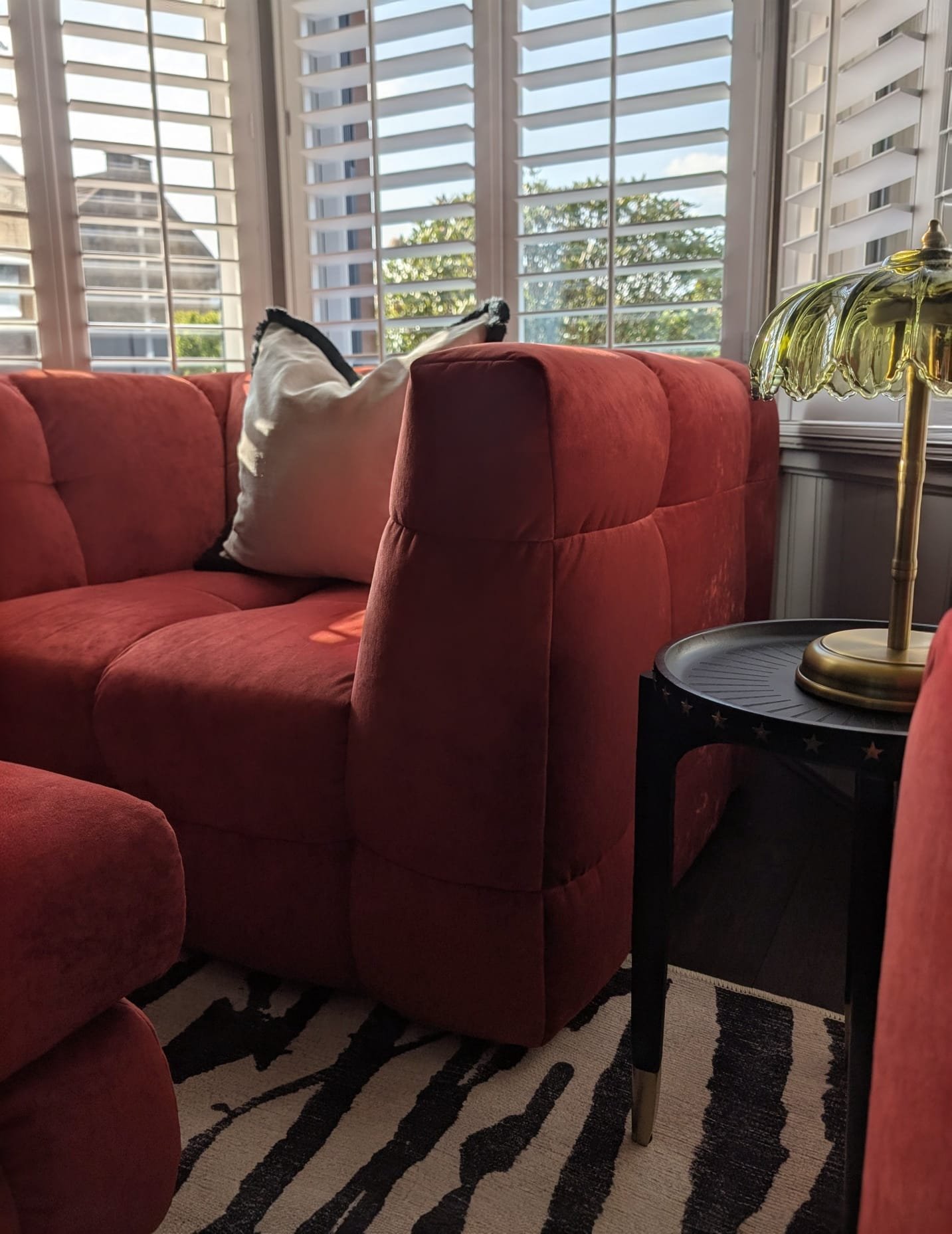 Living room with red velvet sofa, decorative pillow, window with white plantation shutters, black and white striped rug, and a side table with a vintage-style brass lamp.