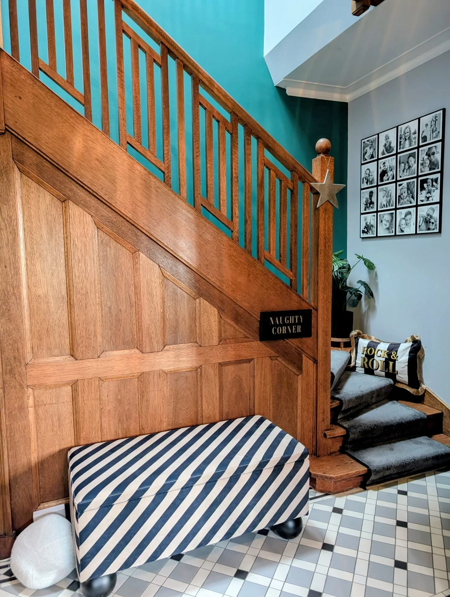 Indoor space with wooden staircase and wall decor. The wall features a collage of black and white photos. There are striped cushions on a bench and pillows labeled "Rock & Roll." A sign reads "Naughty Corner." A potted plant is nearby, and patterned floor tiles are visible.