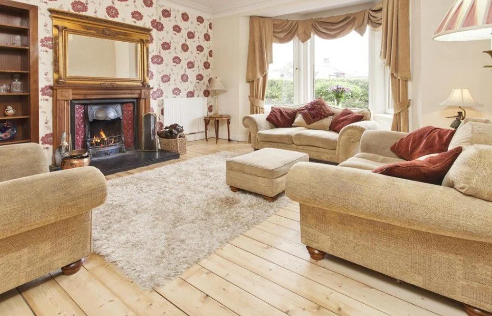Living room with two beige sofas, a matching ottoman, a fireplace with a gold-framed mirror above, a large window with gold curtains, a shaggy beige rug, and wooden floors.