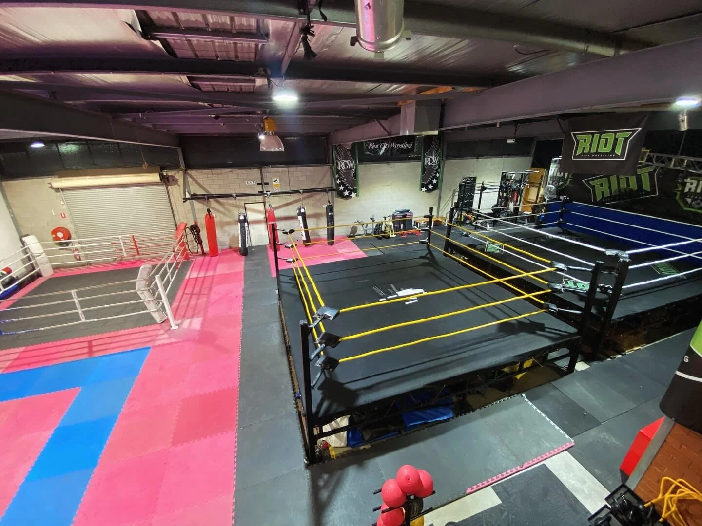 An indoor gym with two wrestling rings and boxing bags, gym equipment, and a pink and blue padded floor area.
