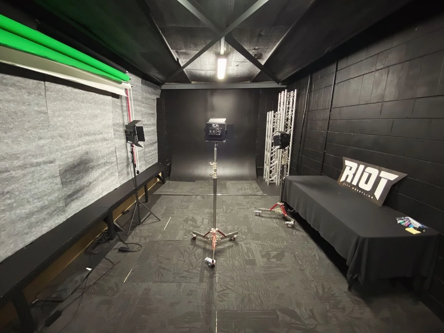 Empty promo studio with a gray and black wall, a black backdrop, studio lights, and a RIOT sign on a table.
