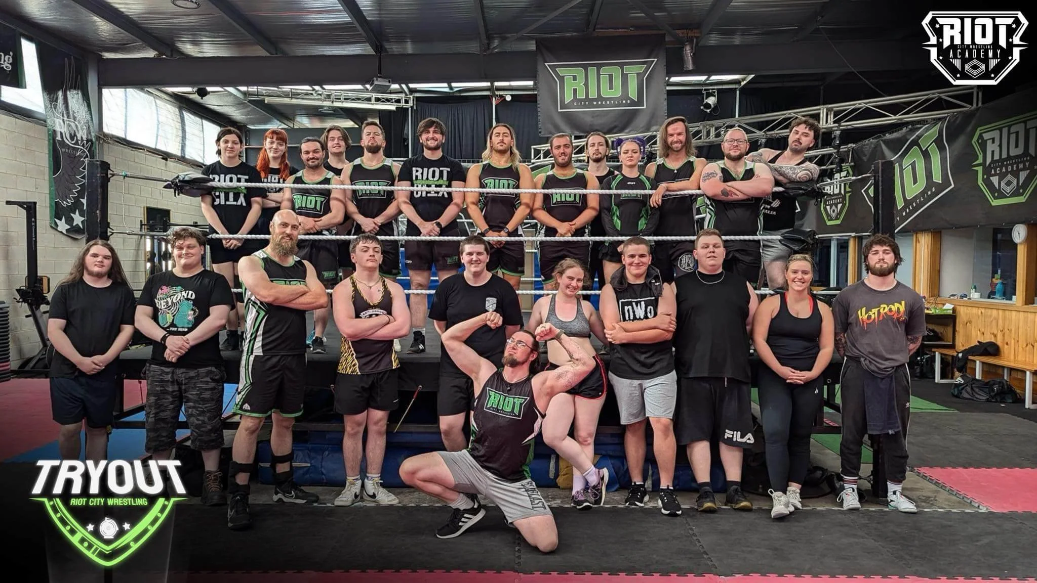 Group photo of a past RCW tryout, with all coaches/active trainees wearing 'Riot City Wrestling' gear, with wrestling rings and banners in the background.