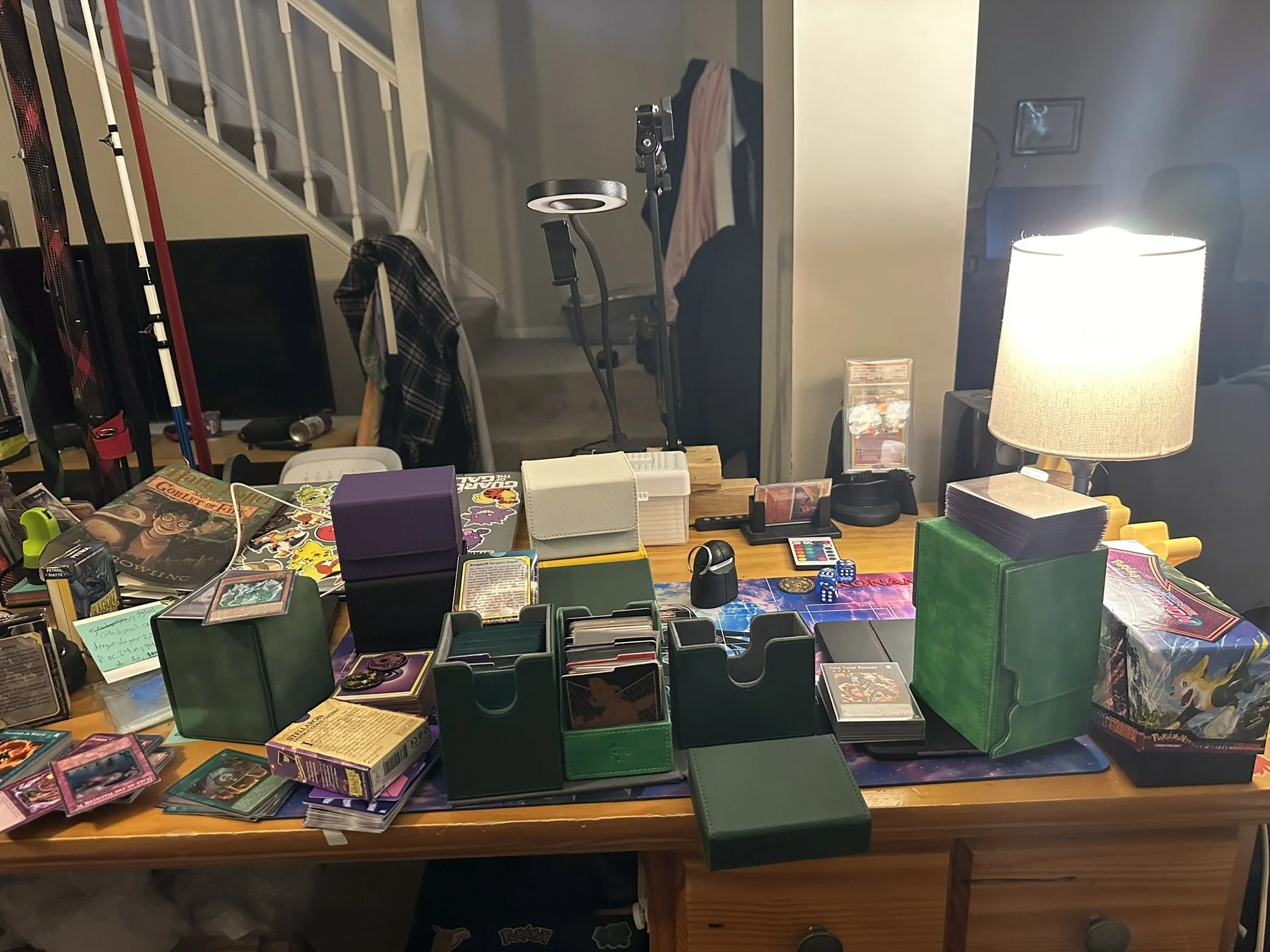 A cluttered wooden desk filled with various trading card game packs, boxes, loose cards, a small deck box, and other gaming accessories. In the background, there is a dimly lit room with a staircase, clothing hanging on the back of a chair, and a lamp illuminating part of the desk.
