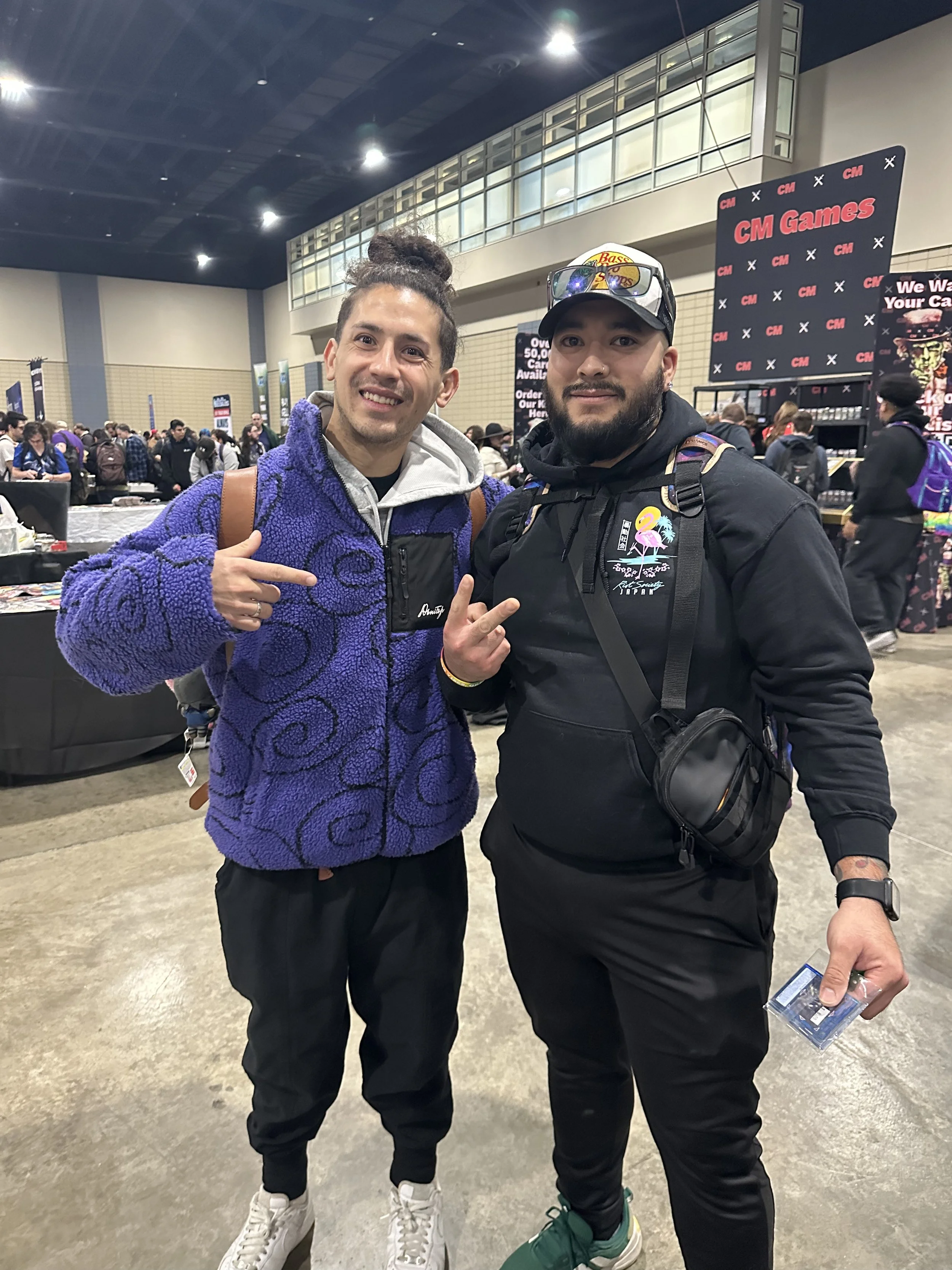 Two men standing in a crowded indoor event, smiling and pointing at each other. One is wearing a purple fleece jacket and has his hair in a bun; the other is dressed in black with a cap, glasses on his head, and holding a clear plastic case.