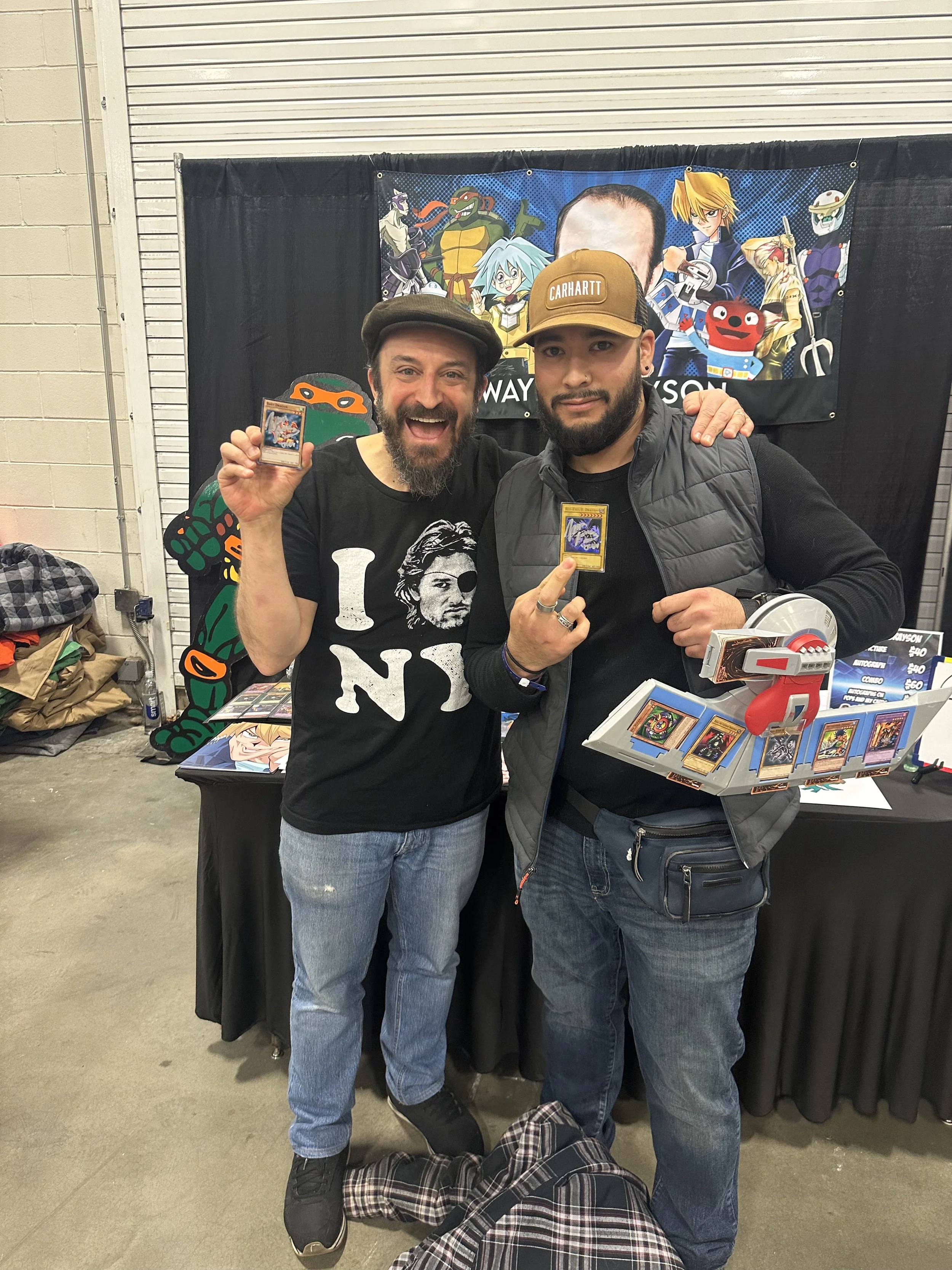 Two men standing together at a convention, holding trading cards, in front of a banner with animated character artwork, with a table displaying more cards and merchandise nearby.
