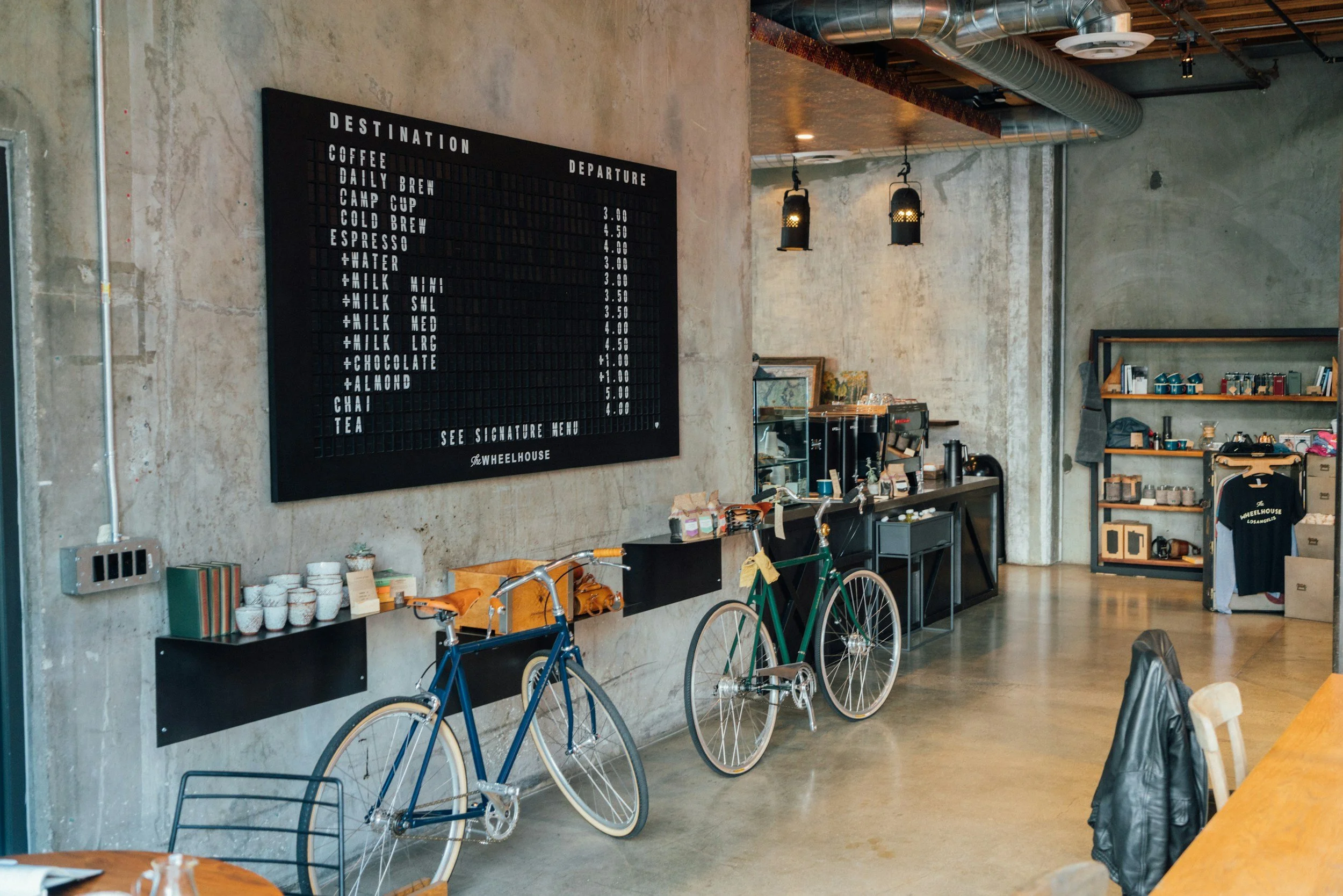 Interior of a coffee shop with a black menu board on the wall listing beverage options and prices, two bicycles parked against the wall, wooden tables and chairs, and shelves with coffee-related items and merchandise.