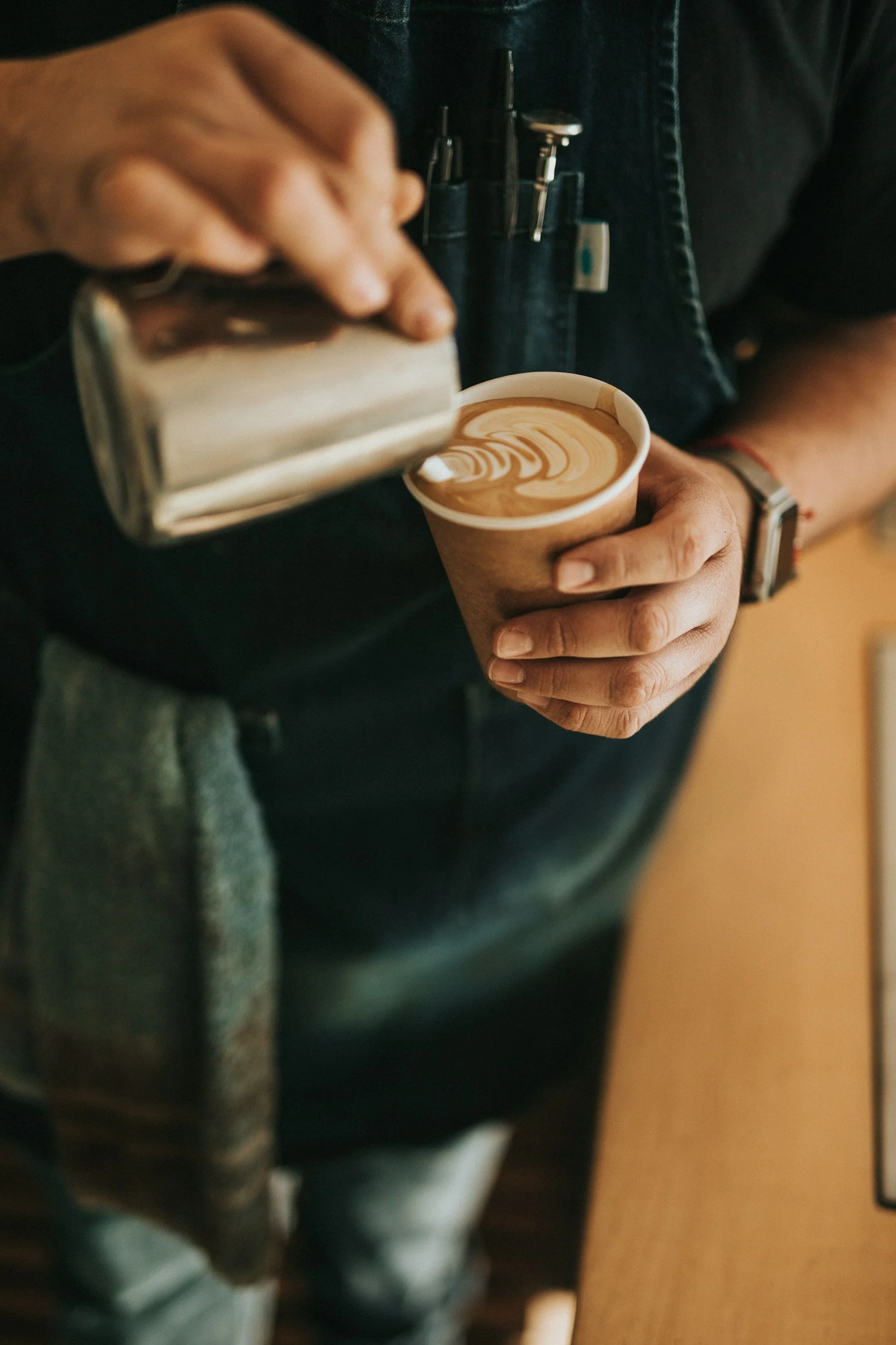 A person in a black shirt and denim apron is pouring steamed milk into a cup of espresso, creating latte art with a heart-shaped pattern.