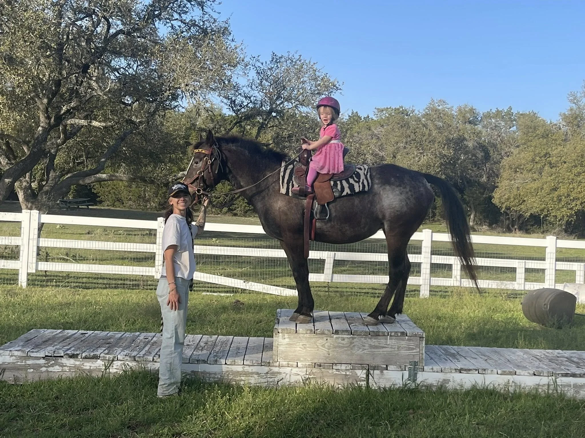 Young girl in a pink helmet sitting on a horse with an instructor at The Gentle Cowgirl in Stockdale, TX — safe, confidence-building horseback riding lessons for young kids near San Antonio