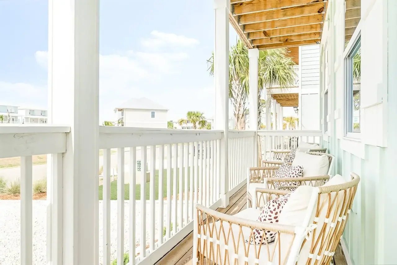A balcony with white railing and seating area, featuring wicker chairs with white cushions and patterned pillows, overlooking a sunny outdoor scene with palm trees and modern houses under a blue sky.
