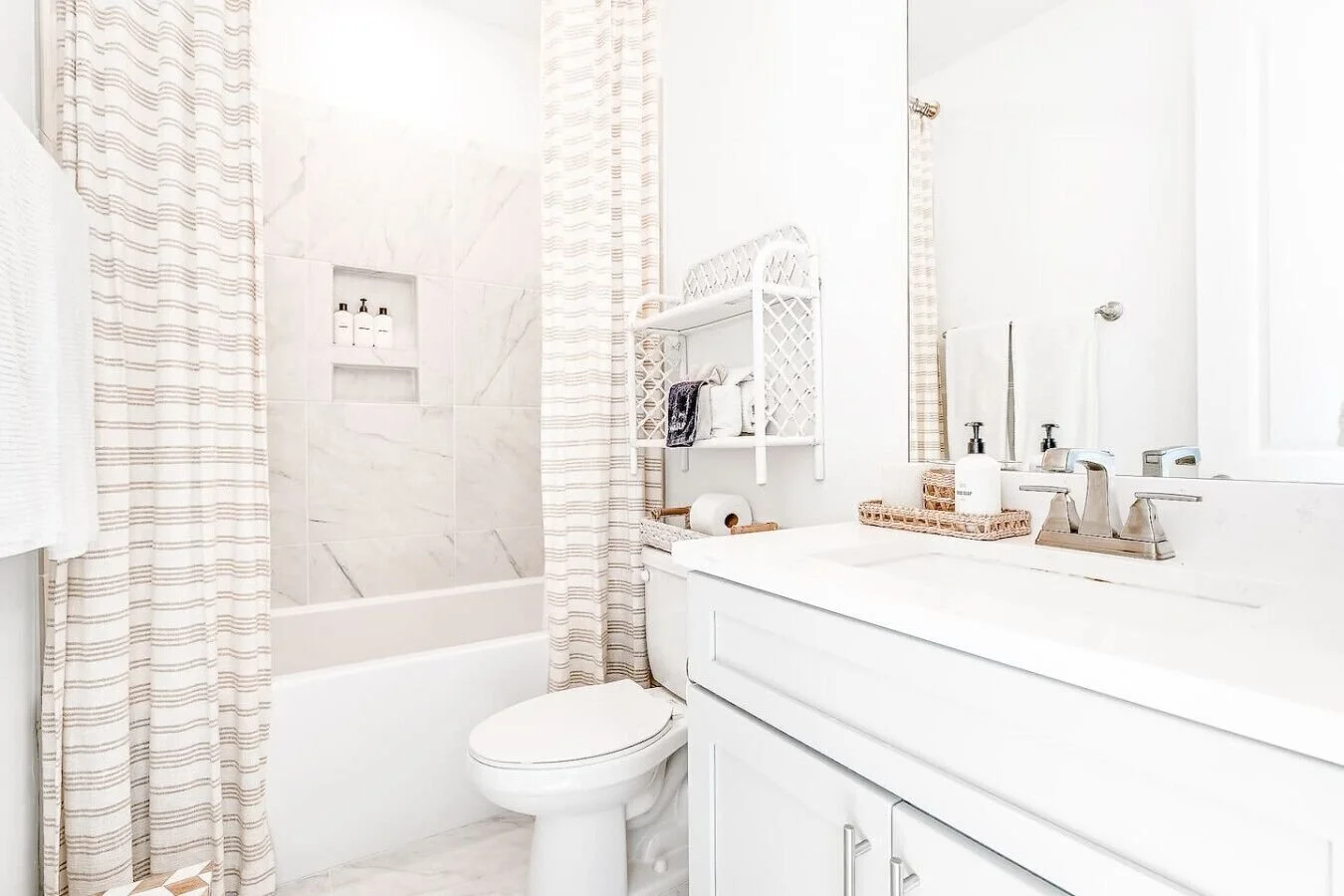Bathroom with a bathtub, shower curtain, toilet, white sink, mirror, towel, soap dispenser, and shelving with various toiletries.
