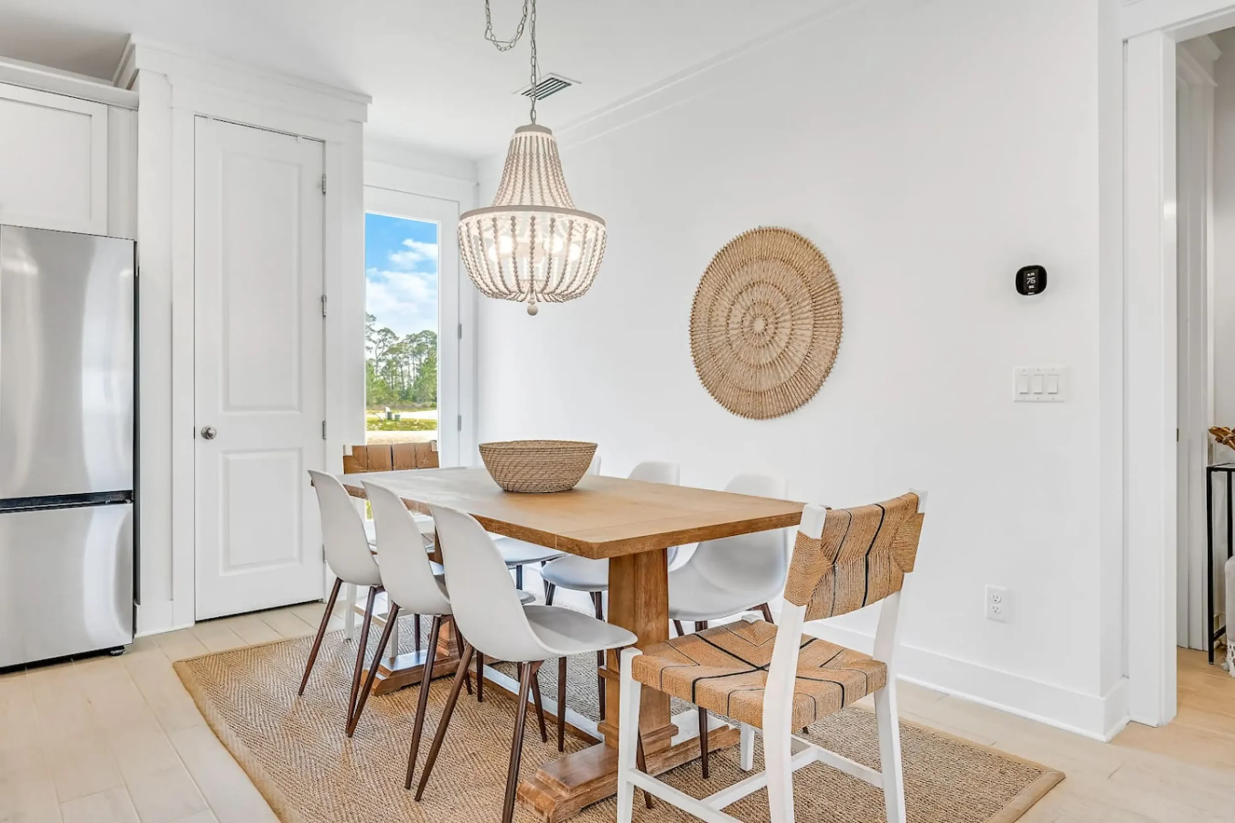 Dining room with a wooden table surrounded by a mix of white and wood chairs, a woven basket centerpiece, a woven wall hanging, and a chandelier, with a door and window showing a view of greenery outside.