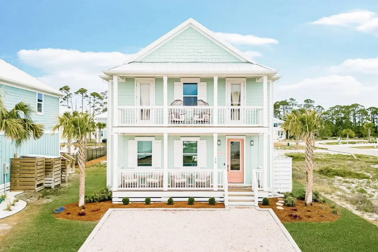 A two-story beach house painted in pastel mint green with white trim, featuring front and upper balconies, with two wicker chairs on the upper balcony, a coral-colored front door, and a small front porch with stairs. Vacation rental Cape San Blas