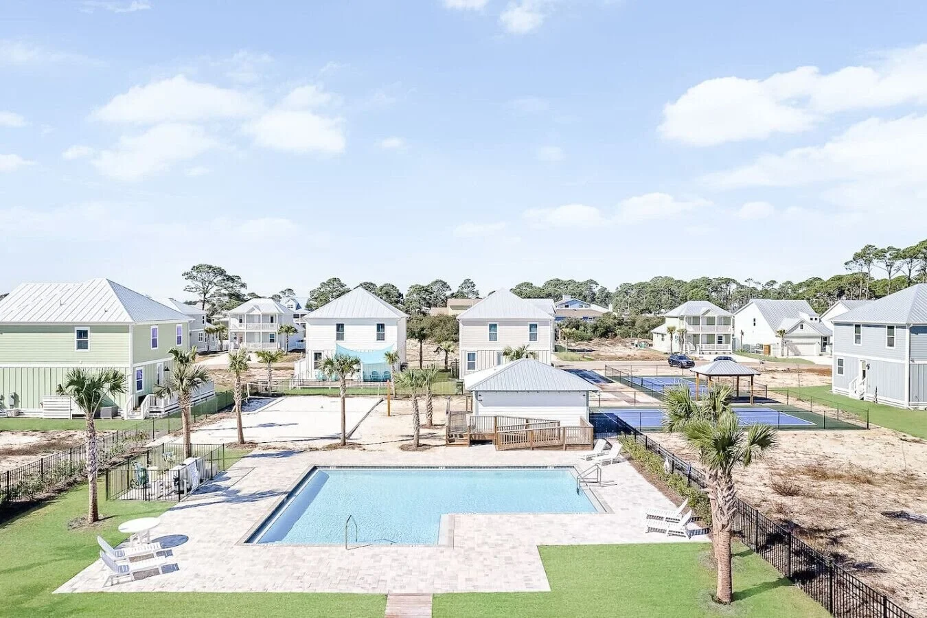 A swimming pool area with lounge chairs and a small round table, surrounded by a fence with palm trees, in a residential neighborhood with multiple houses and tennis courts visible in the background.