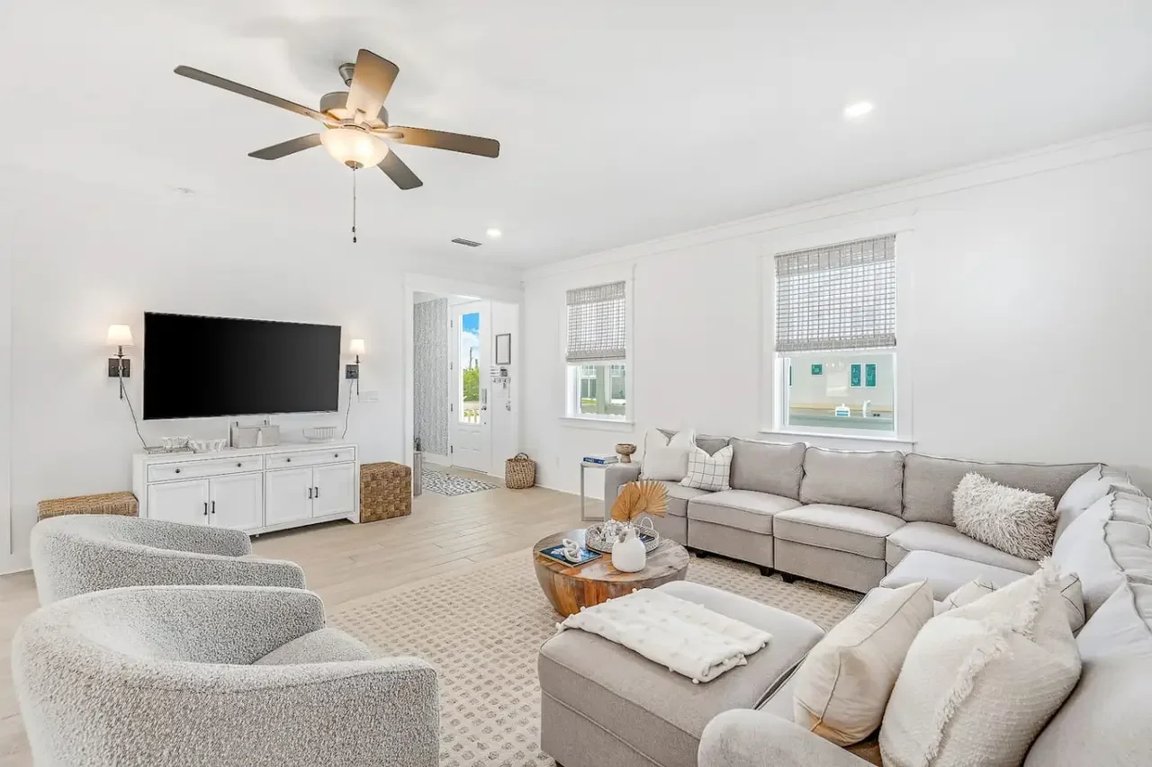 Living room with a large sectional sofa, a flat-screen TV on a white console, two armchairs, a round wooden coffee table, and three windows with bamboo shades. Walls are white, with a ceiling fan and wall sconces.