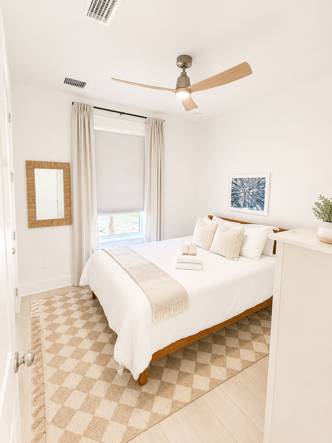 Bright bedroom with white walls, a wooden bed with white bedding and beige pillows, a bedside table with a plant, a window with white curtains, a framed artwork, and a beige diamond-patterned rug.