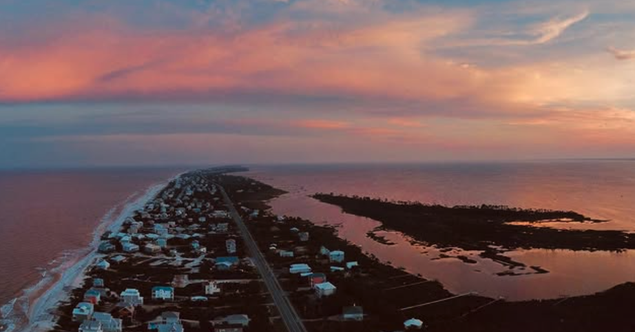 An aerial view of a coastal area during sunset, with a long road running alongside residential houses and a body of water on either side, one with calmer waters and the other with wave activity.