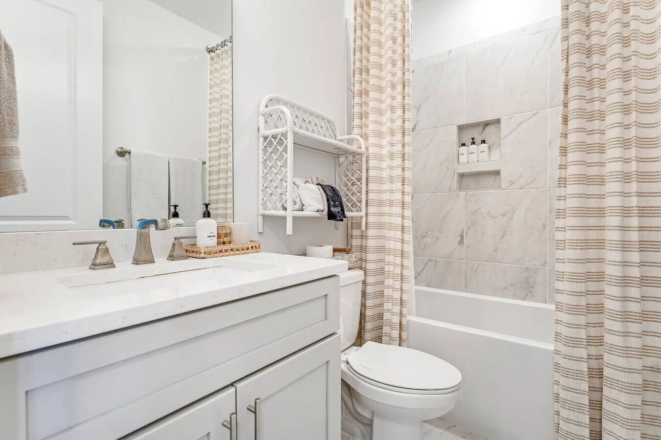 Bathroom with white vanity, marble countertop, toothbrush, soap dispenser, towel rack, mirror, toilet, shower with marble tiles, built-in shampoo shelf, and striped shower curtain.