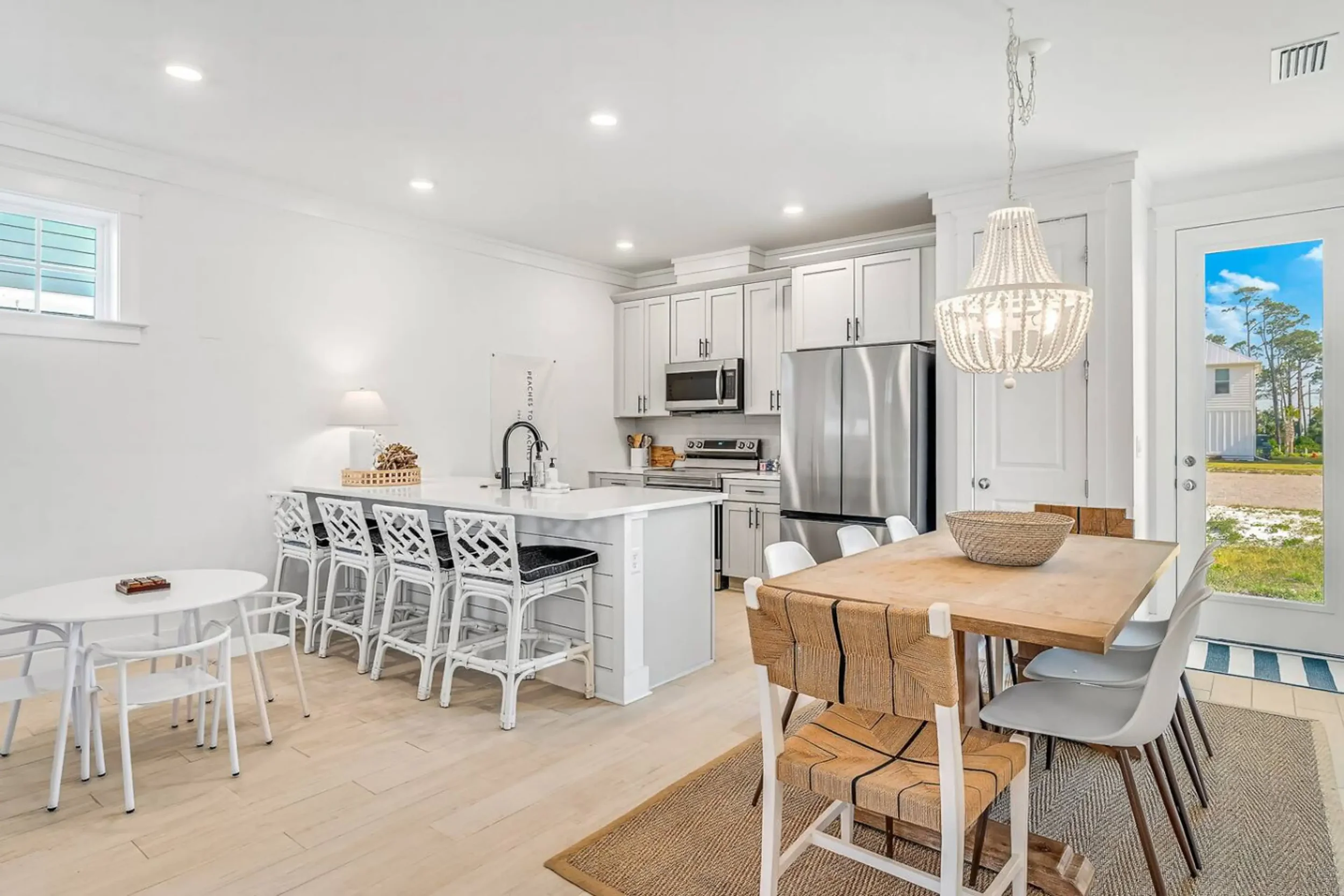 Bright, modern kitchen and dining area with white cabinets, stainless steel appliances, and a variety of chairs around a wooden dining table. A large window overlooking a yard, with a white pendant light hanging from the ceiling.