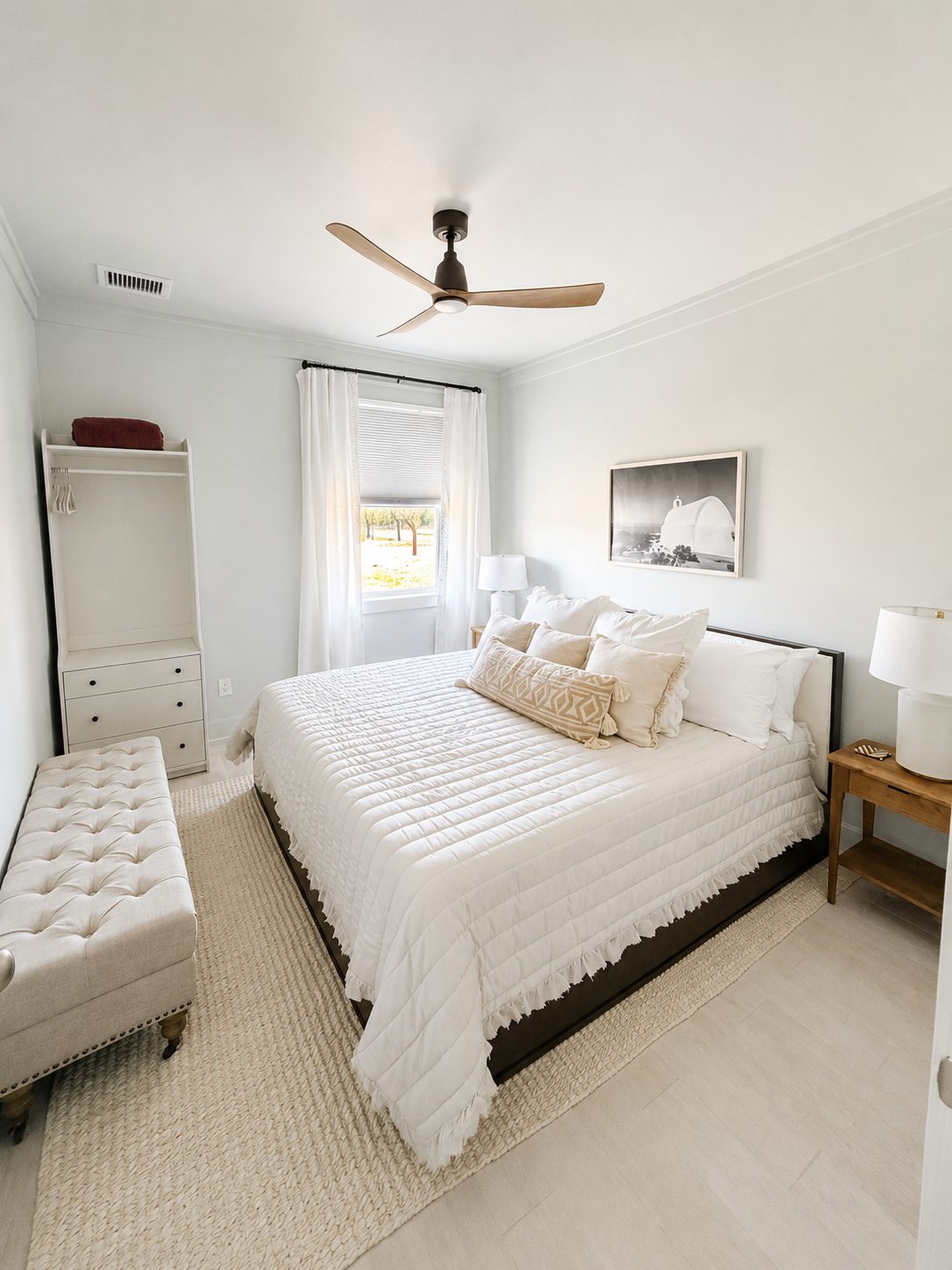 A minimalist bedroom with a white bed, beige and white pillows, matching bedside lamps, a wooden nightstand, a window with white curtains, a framed black-and-white photograph, a tufted bench at the foot of the bed, and a ceiling fan with wooden blades.