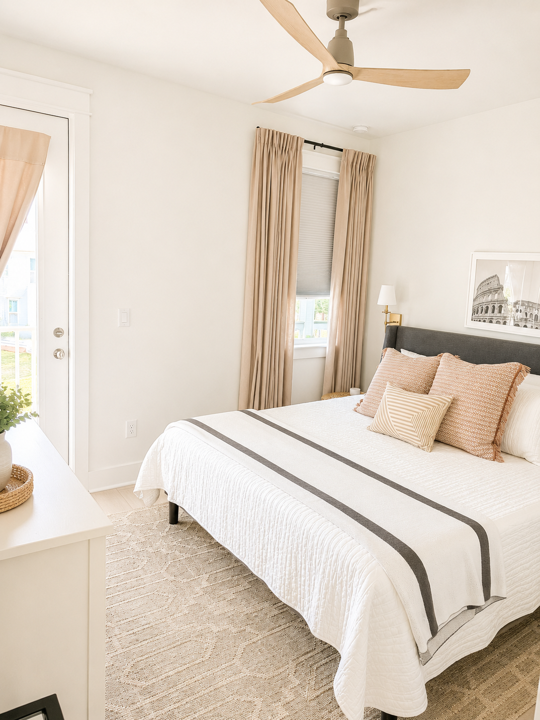 A bright bedroom with a white bed, beige curtains, a ceiling fan, and wall art of a famous landmark.