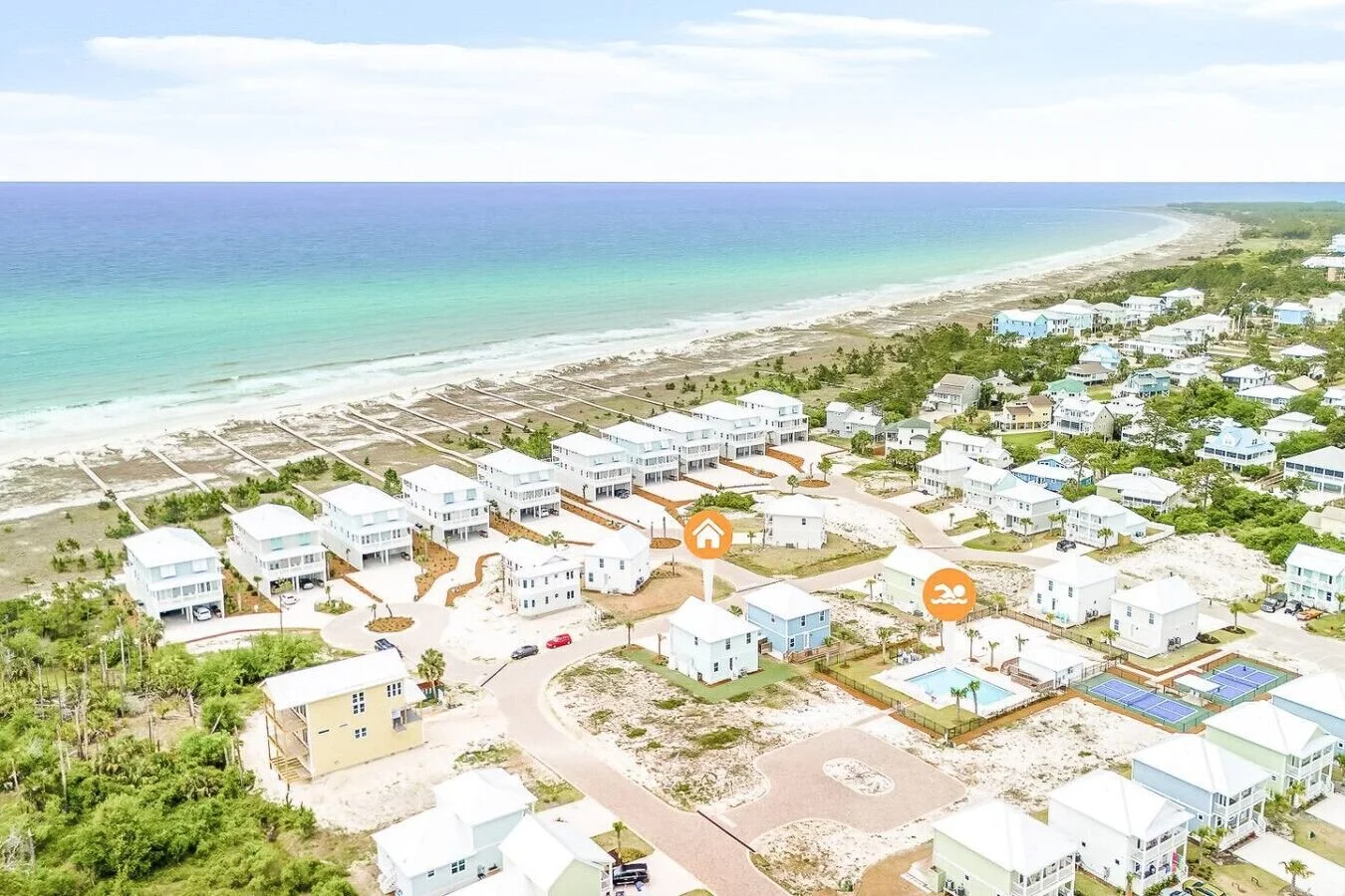 Aerial view of a beachside neighborhood with houses near the ocean, a swimming pool, tennis courts, and lush greenery.