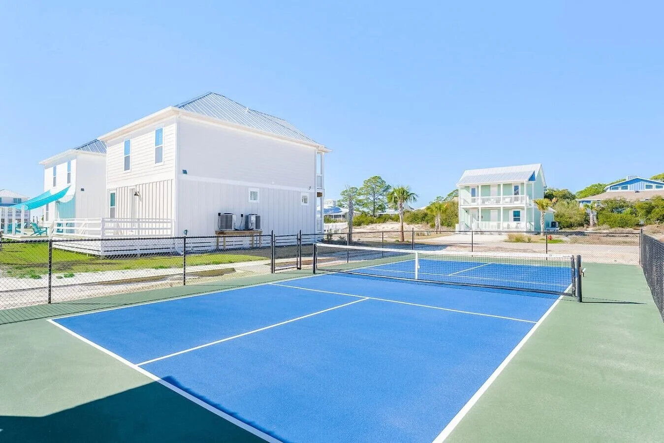 A tennis court with a blue surface in a residential neighborhood under clear sky. Houses with white and light blue exteriors are in the background, with palm trees and greenery around.