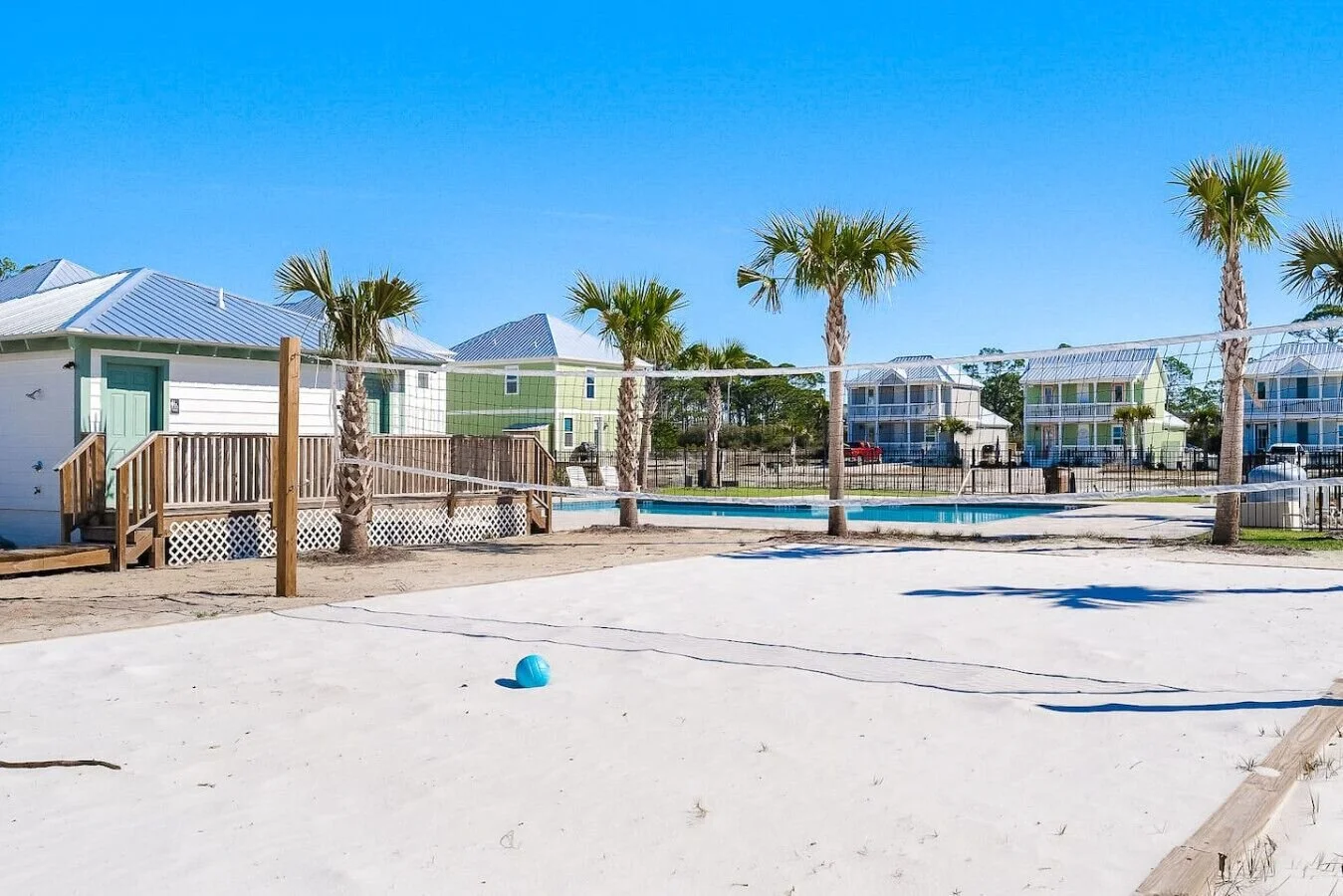 Empty volleyball court with palm trees, a small building, swimming pool, and colorful houses in the background under a clear blue sky.