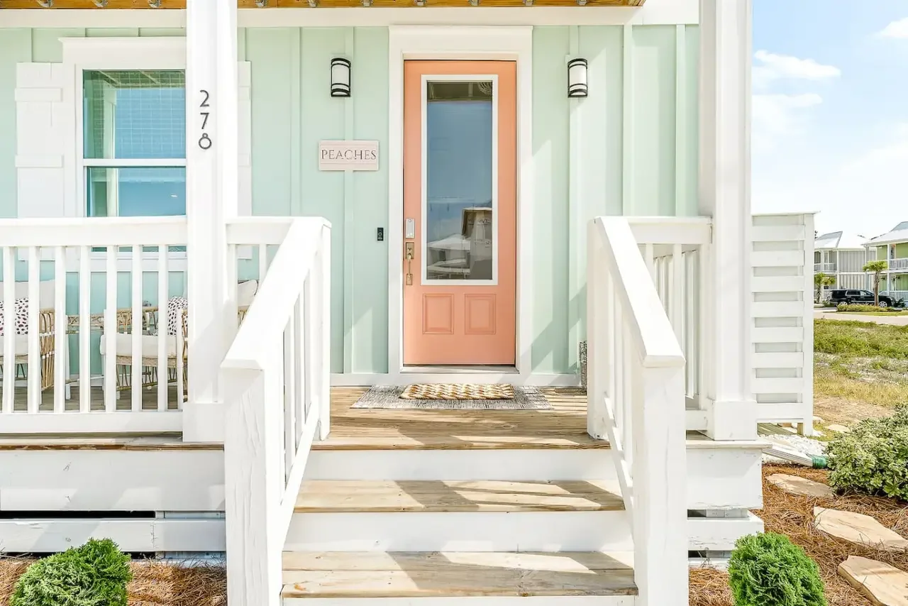 Front porch of a pastel-colored house with a pink front door, white railings, and outdoor lighting fixtures.