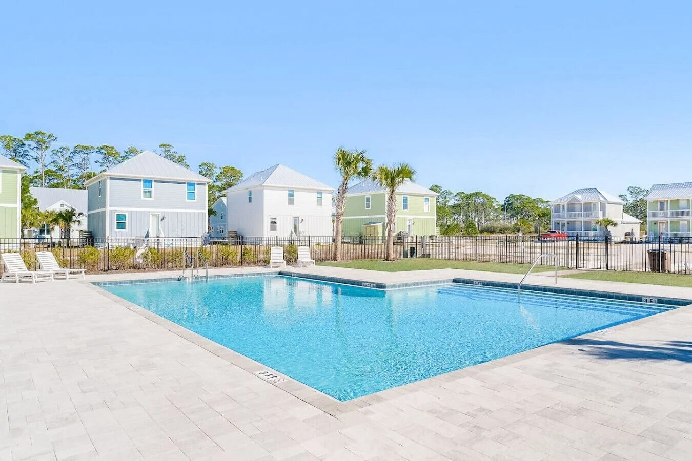 Community swimming pool with lounge chairs, surrounded by colorful houses with palm trees and a clear blue sky.