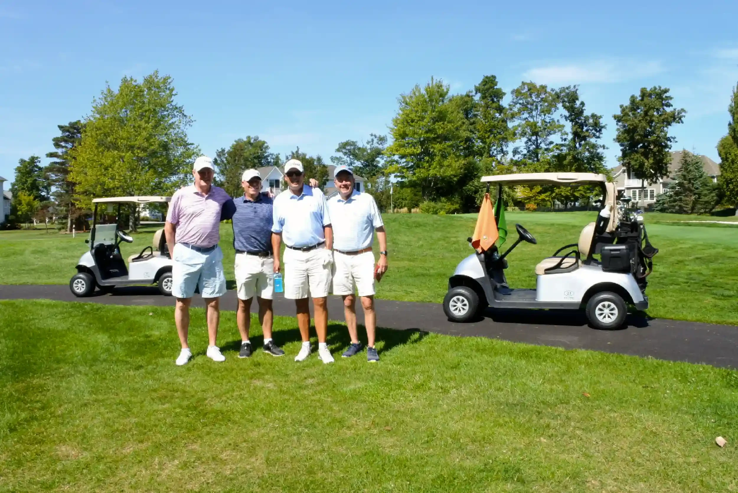 Four men standing together on a golf course, smiling, with two golf carts behind them and green trees in the background.