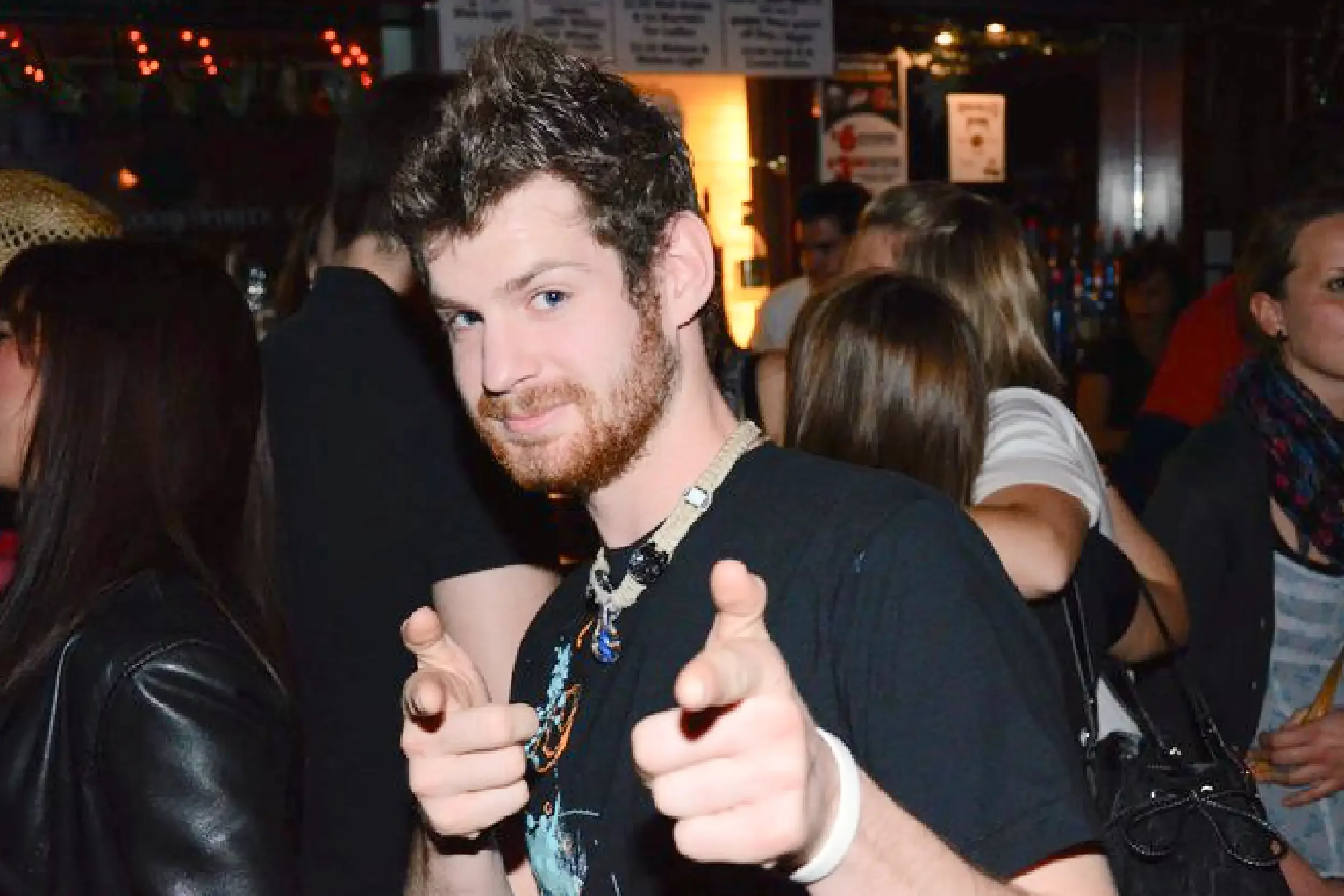 Young man with curly hair and a beard pointing towards the camera, surrounded by people in a lively, dimly lit indoor setting.
