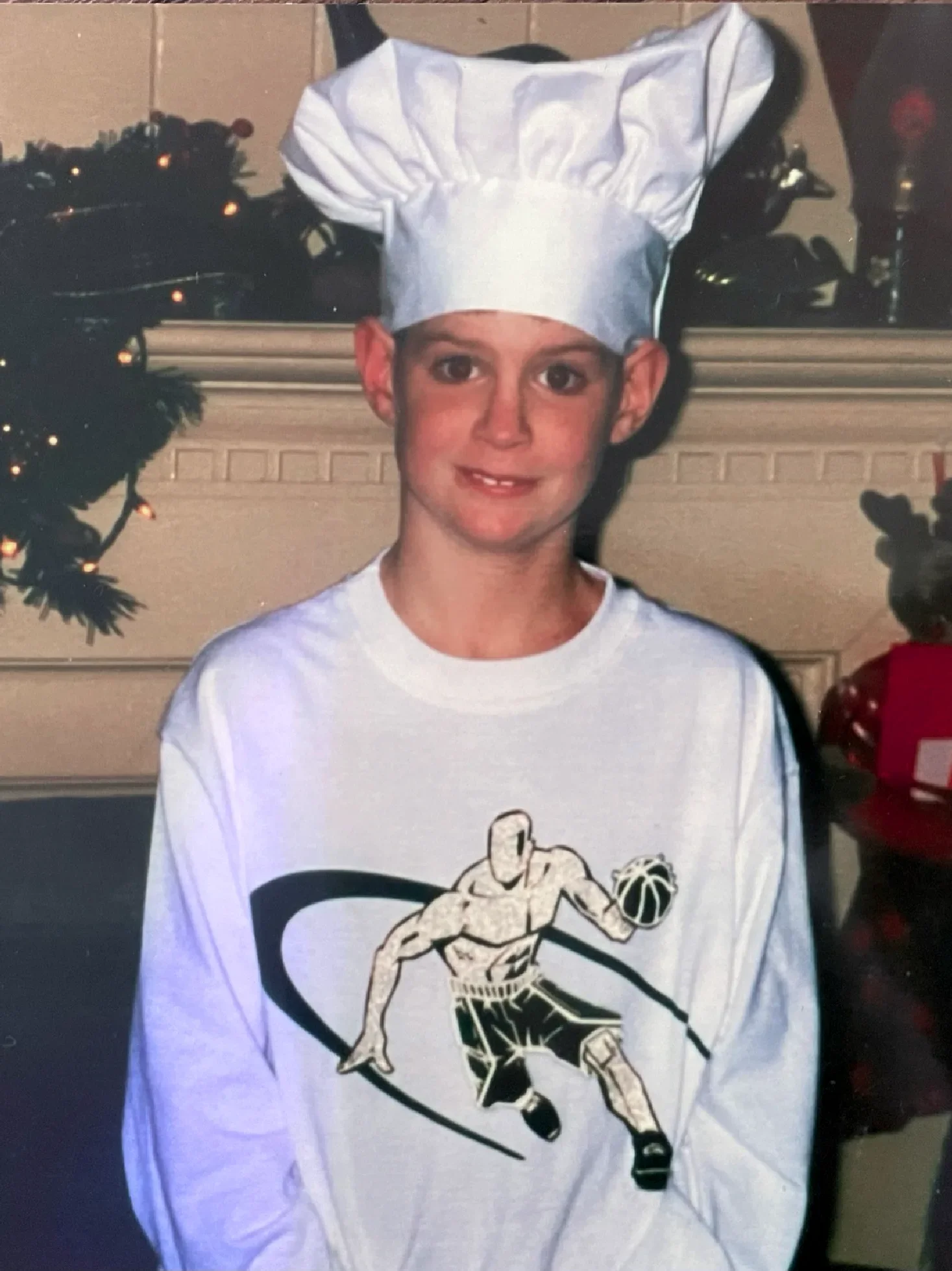 A young person wearing a white hockey jersey with a black and white illustration of a hockey player on the front, and a white chef's hat. They are standing indoors with a decorated Christmas tree visible in the background.