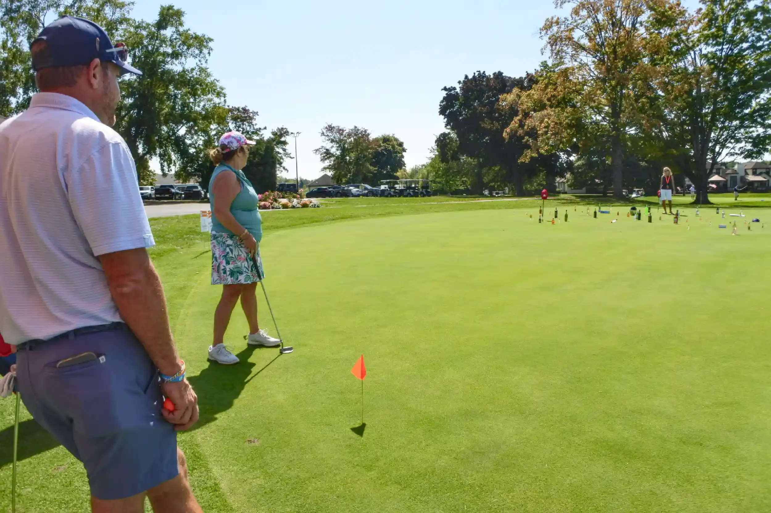Three people on a golf course practicing putting. Two adults are in the foreground, and a child is in the background near the putting green. The scene takes place on a sunny day with trees and parked cars visible in the distance.