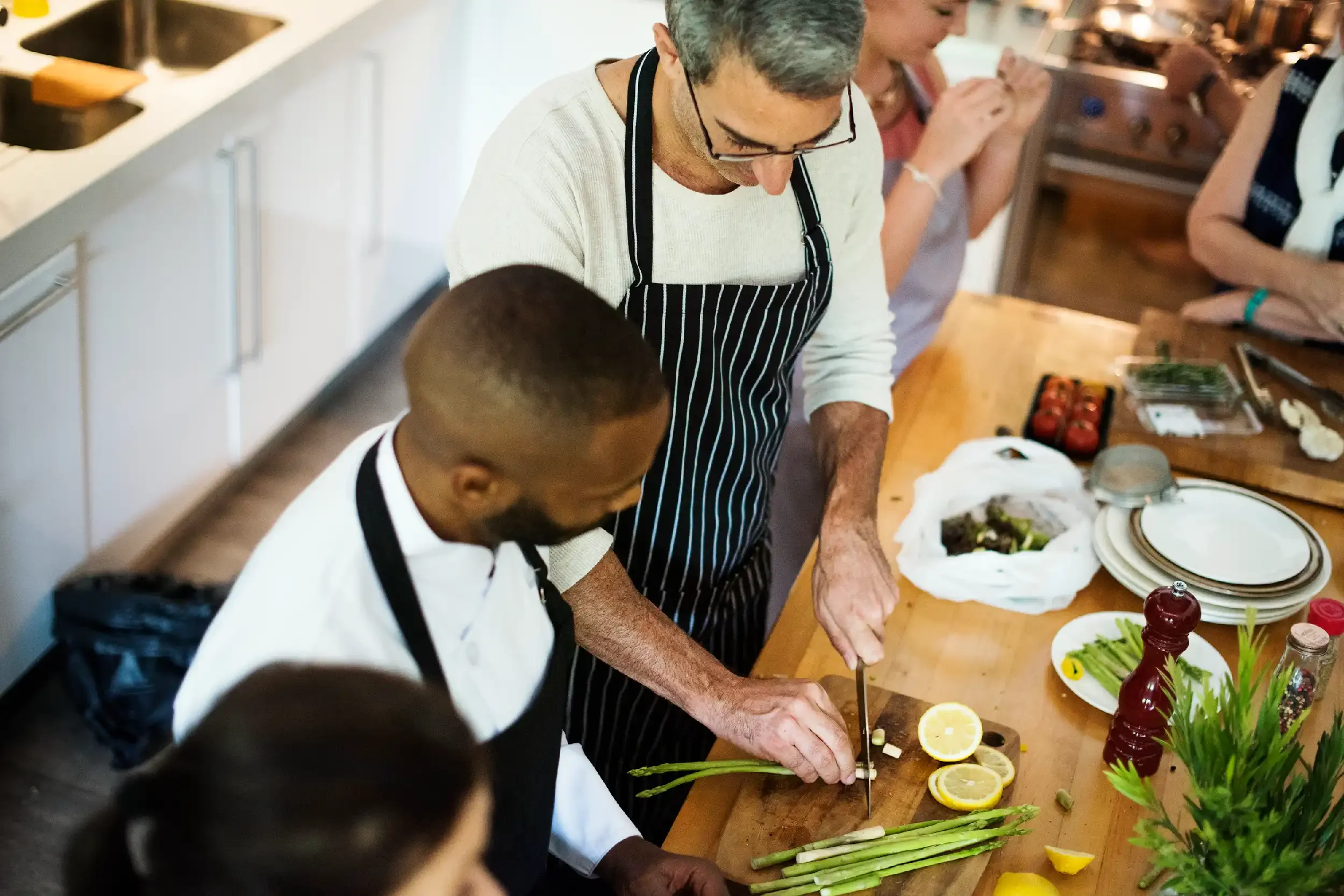 People gathered around a kitchen counter preparing food, with lemon slices, asparagus, and various ingredients on the table.