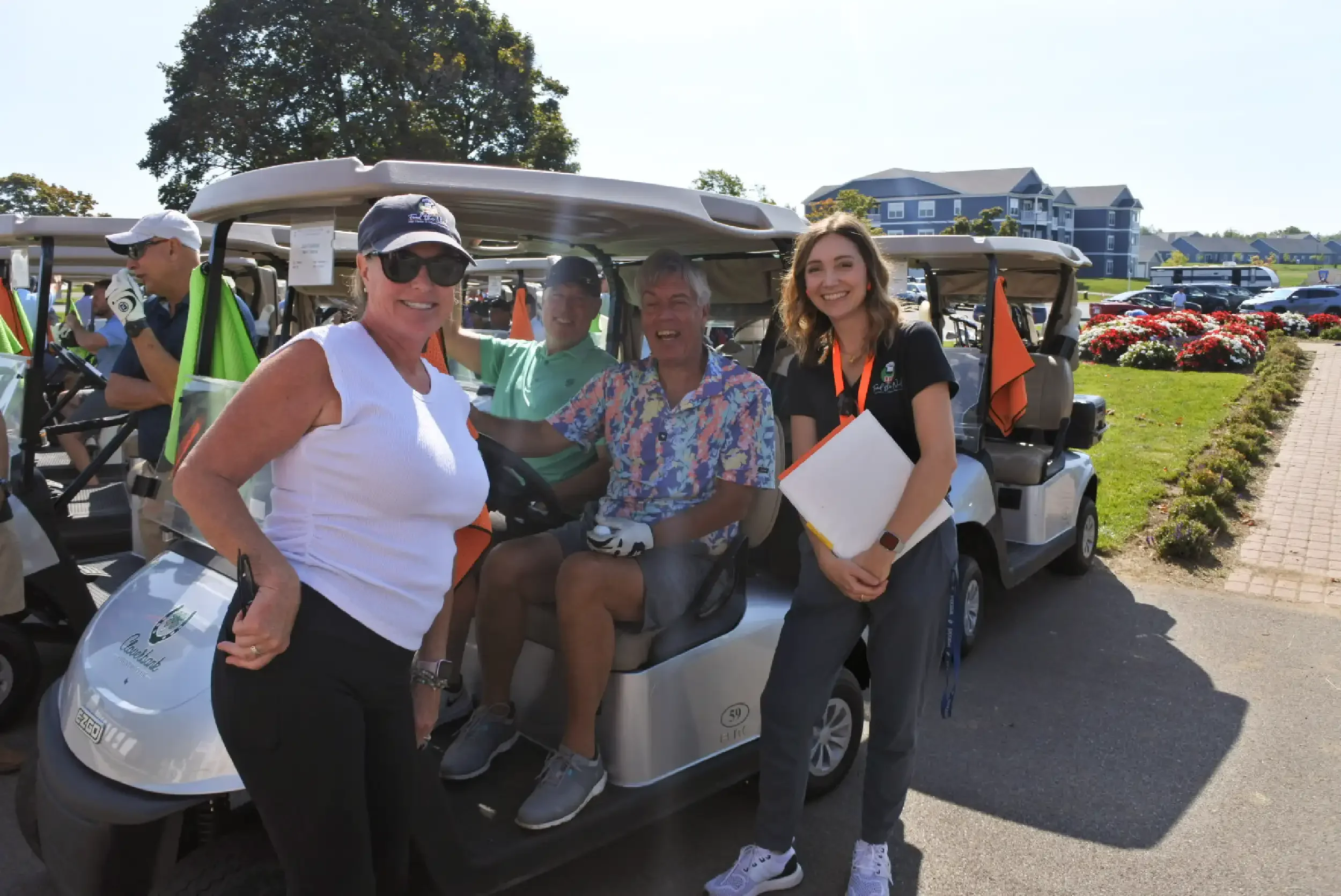 Group of people smiling and standing near golf carts on a golf course with flowers and buildings in the background on a sunny day.