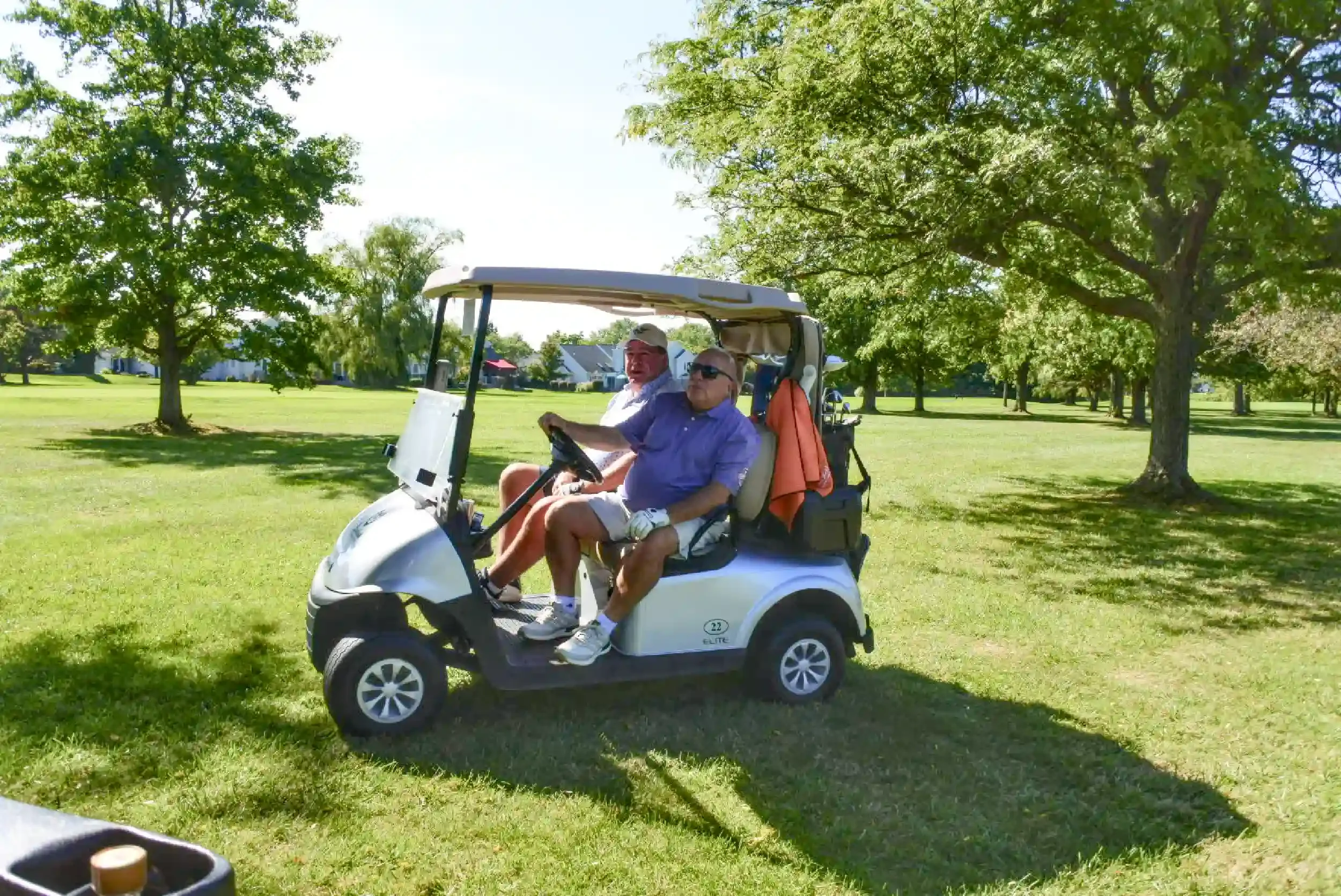 Two older men sitting in a white golf cart on a sunny golf course, surrounded by green grass and large leafy trees.
